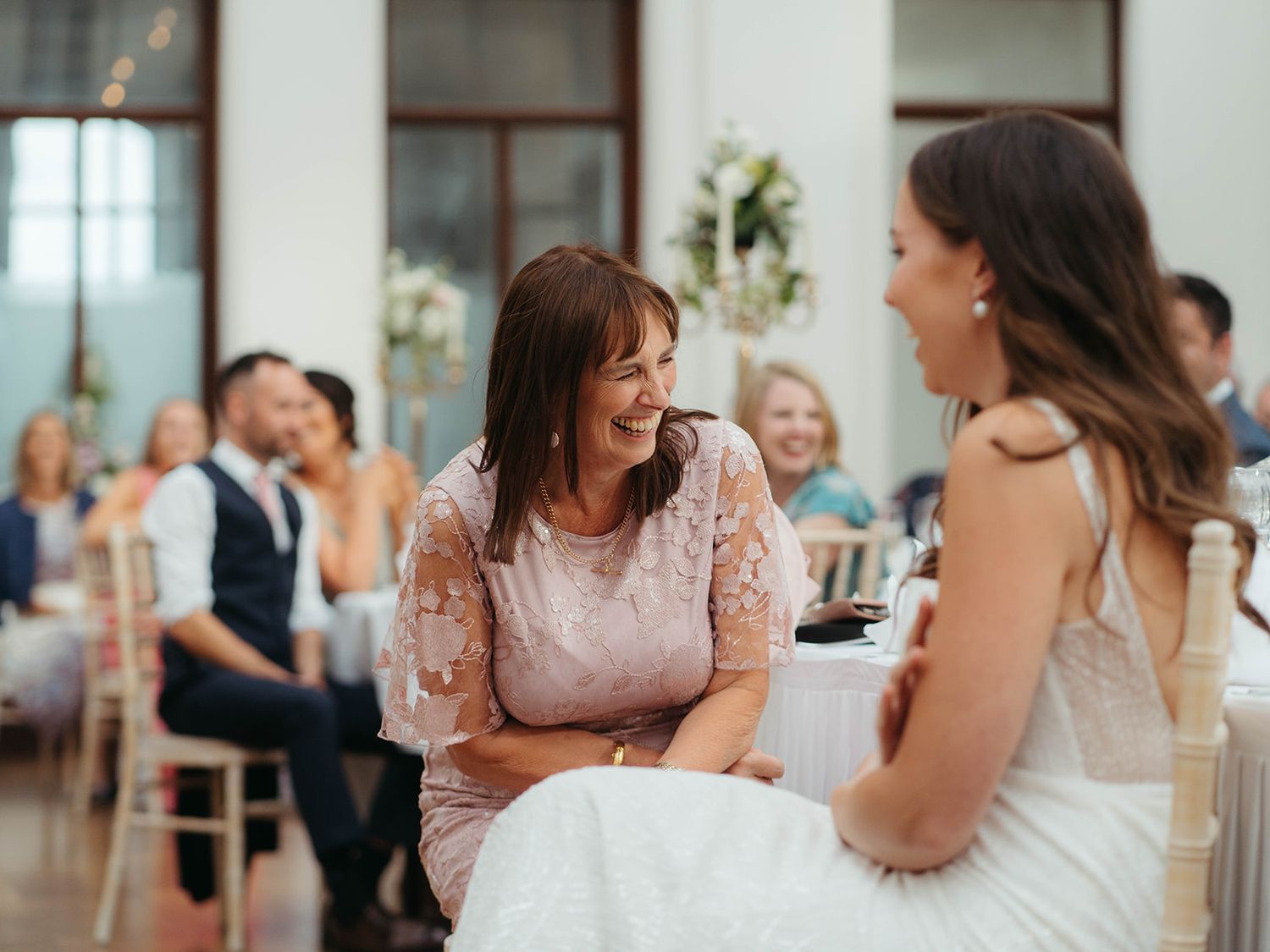 Two wedding guests share a joyful conversation during the reception in an elegant venue with natural lighting.