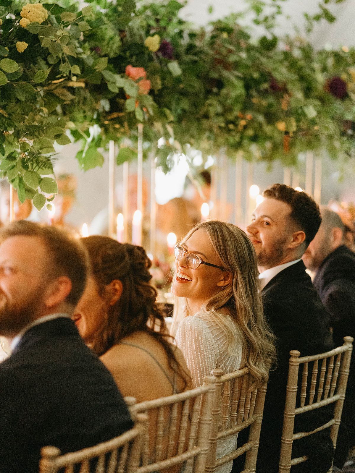 Wedding reception guests seated at candlelit tables under lush floral ceiling decor creating an intimate atmosphere.