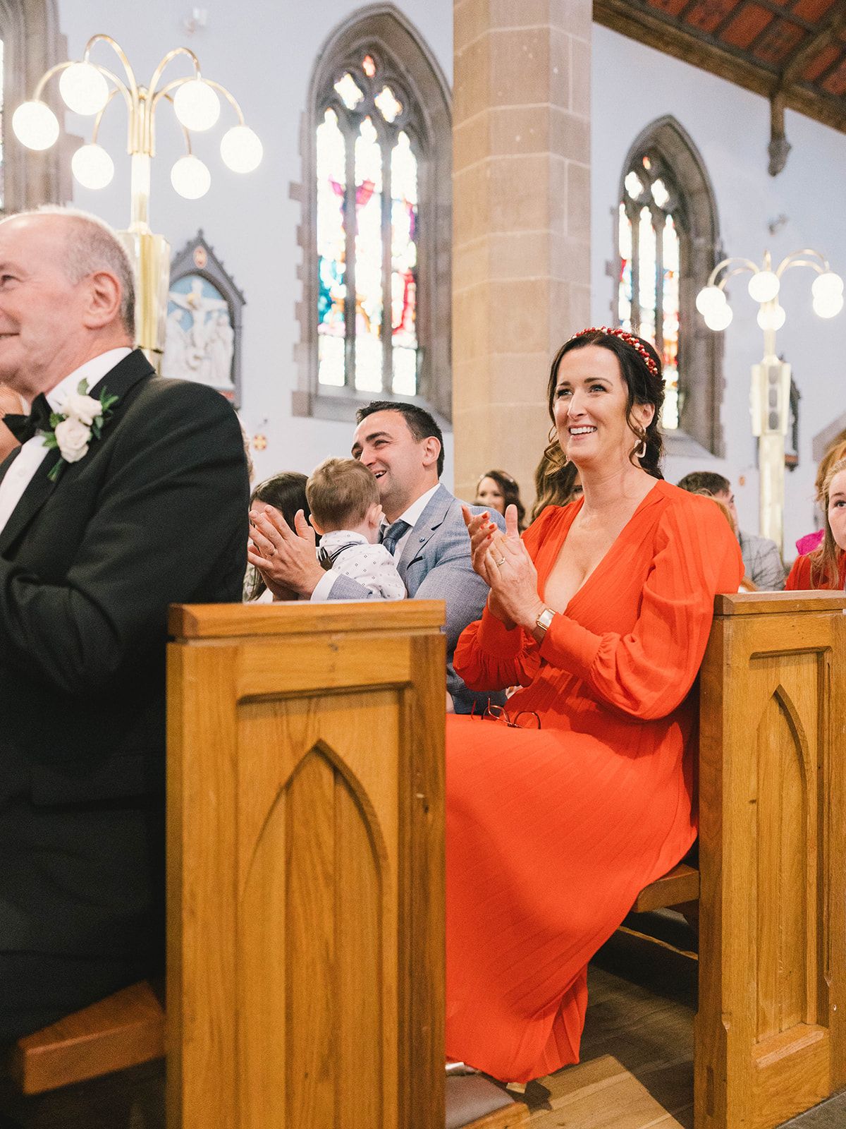 Wedding guests seated in wooden church pews smile during a ceremony with Gothic stained glass windows in the background.