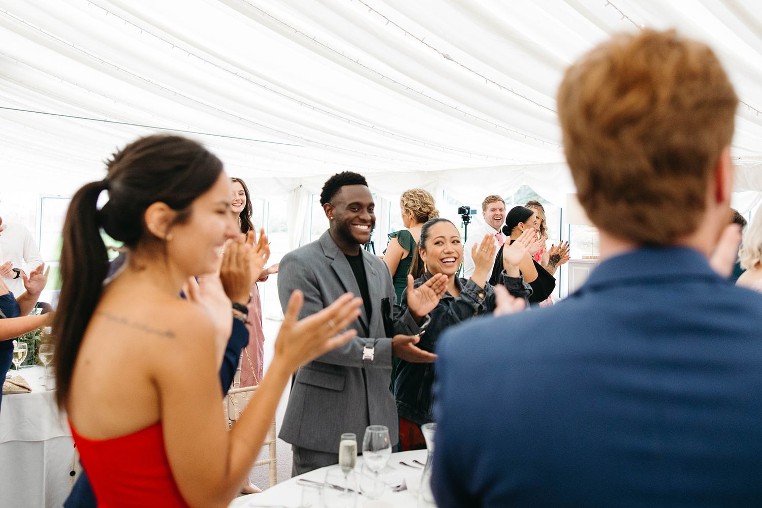 A lively group of wedding guests react with laughter and excitement during a celebration in a white tent reception venue.