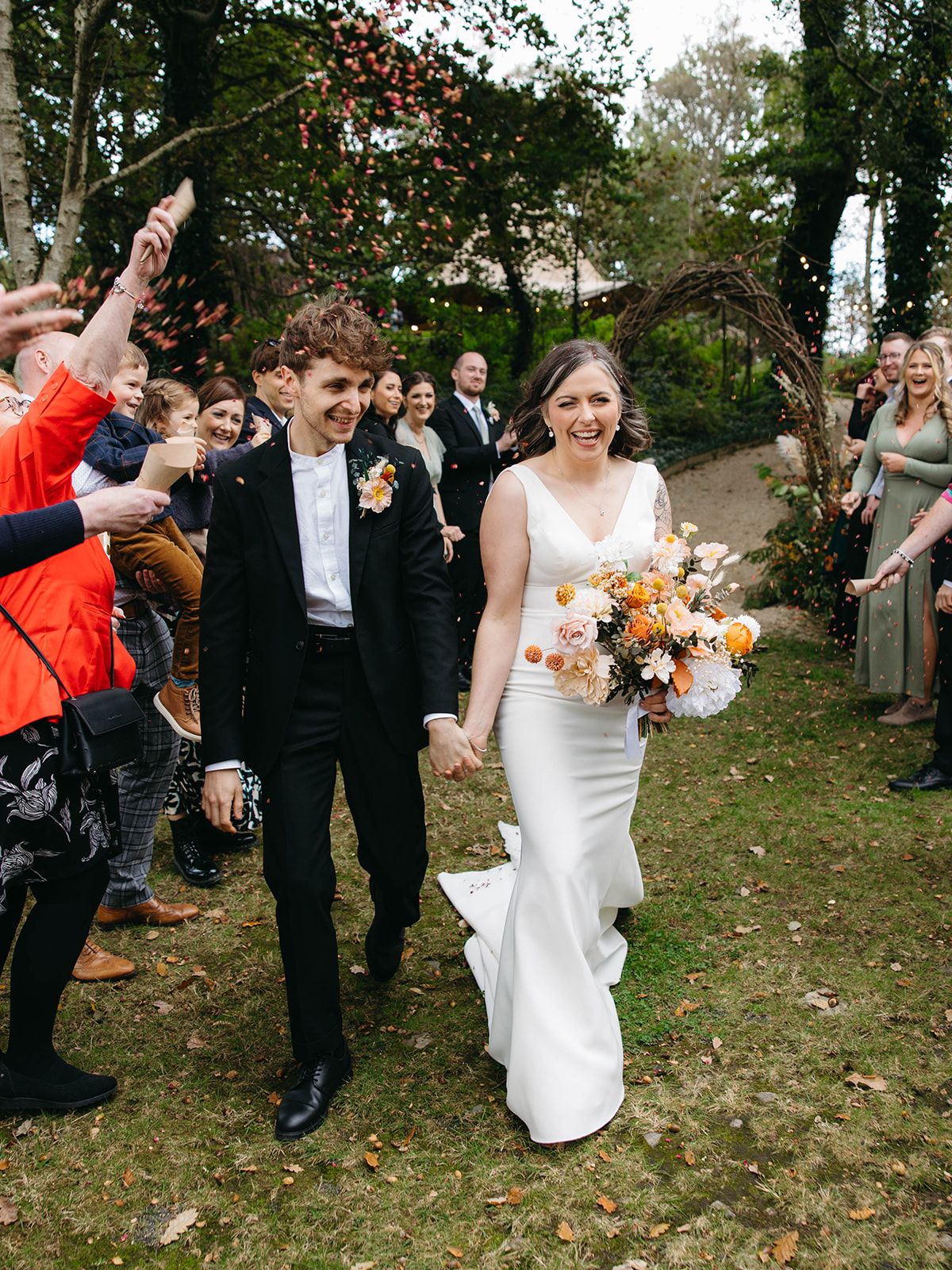 Couple walks together during autumn wedding ceremony beneath rustic arch with guests celebrating outdoors.