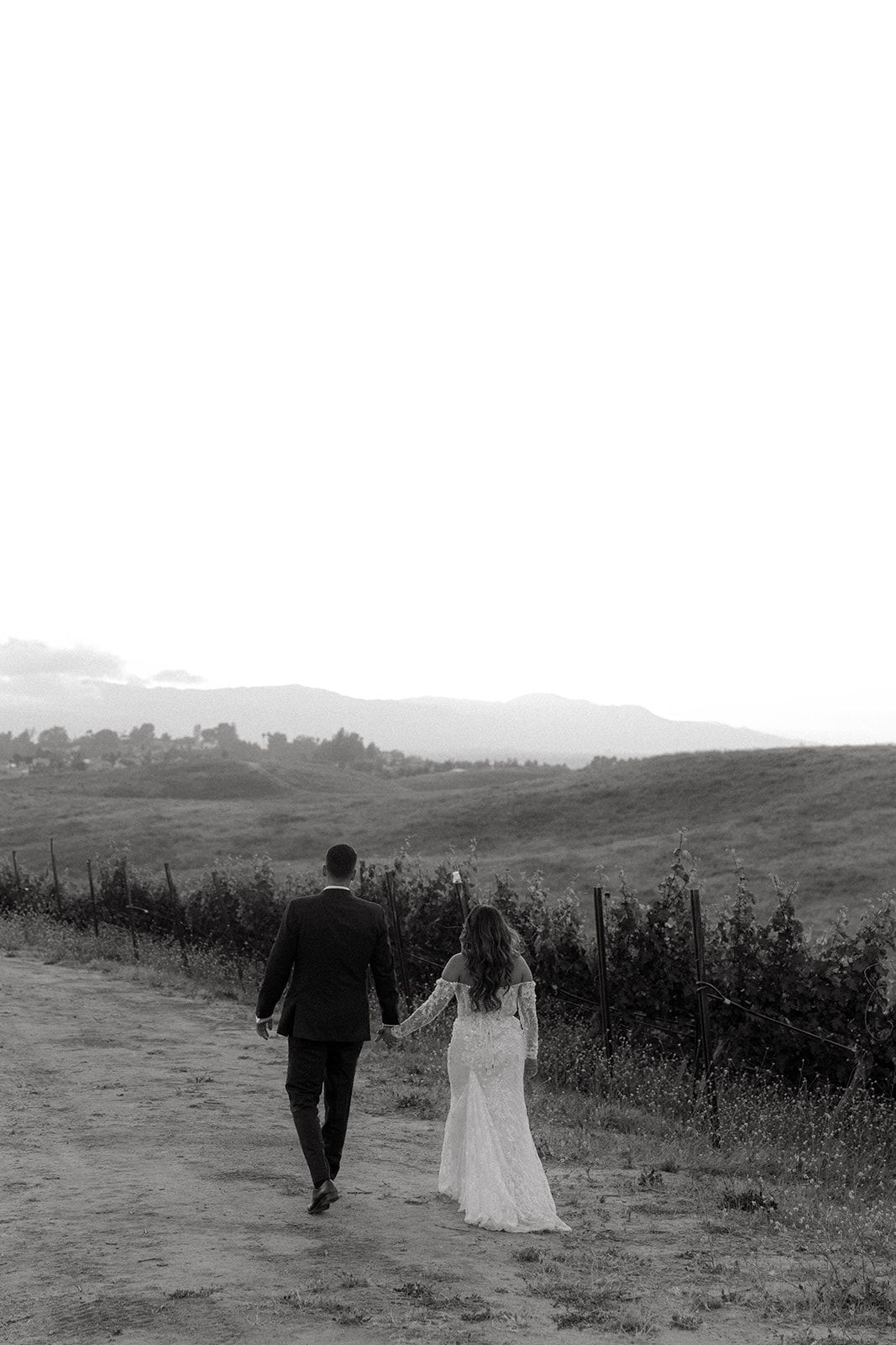 A newlywed couple walks hand in hand along a hilltop path surrounded by vineyard landscapes at sunset.