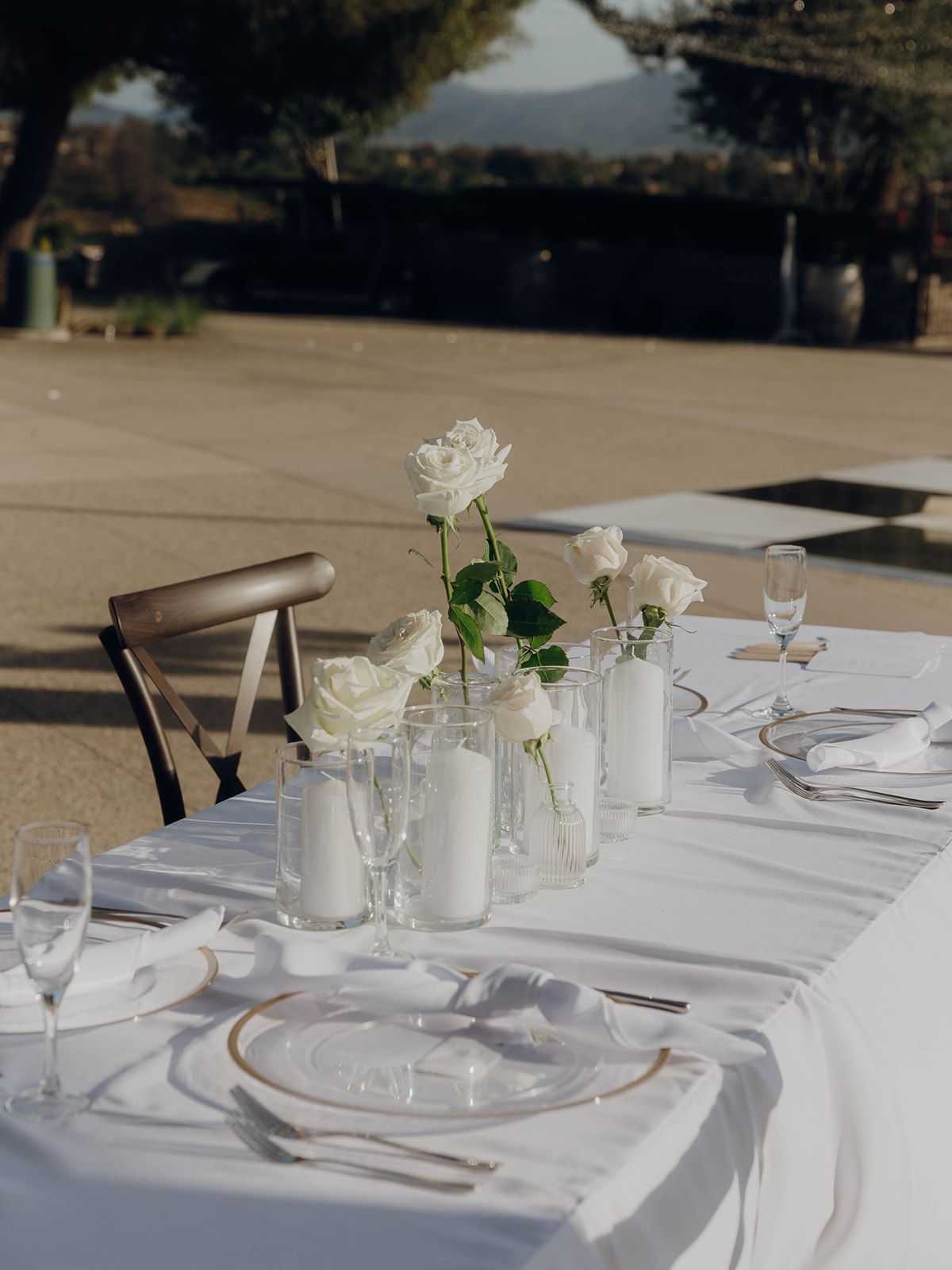 White roses in vases adorn an elegant outdoor table setting with crisp linens and place settings for a formal dining event.