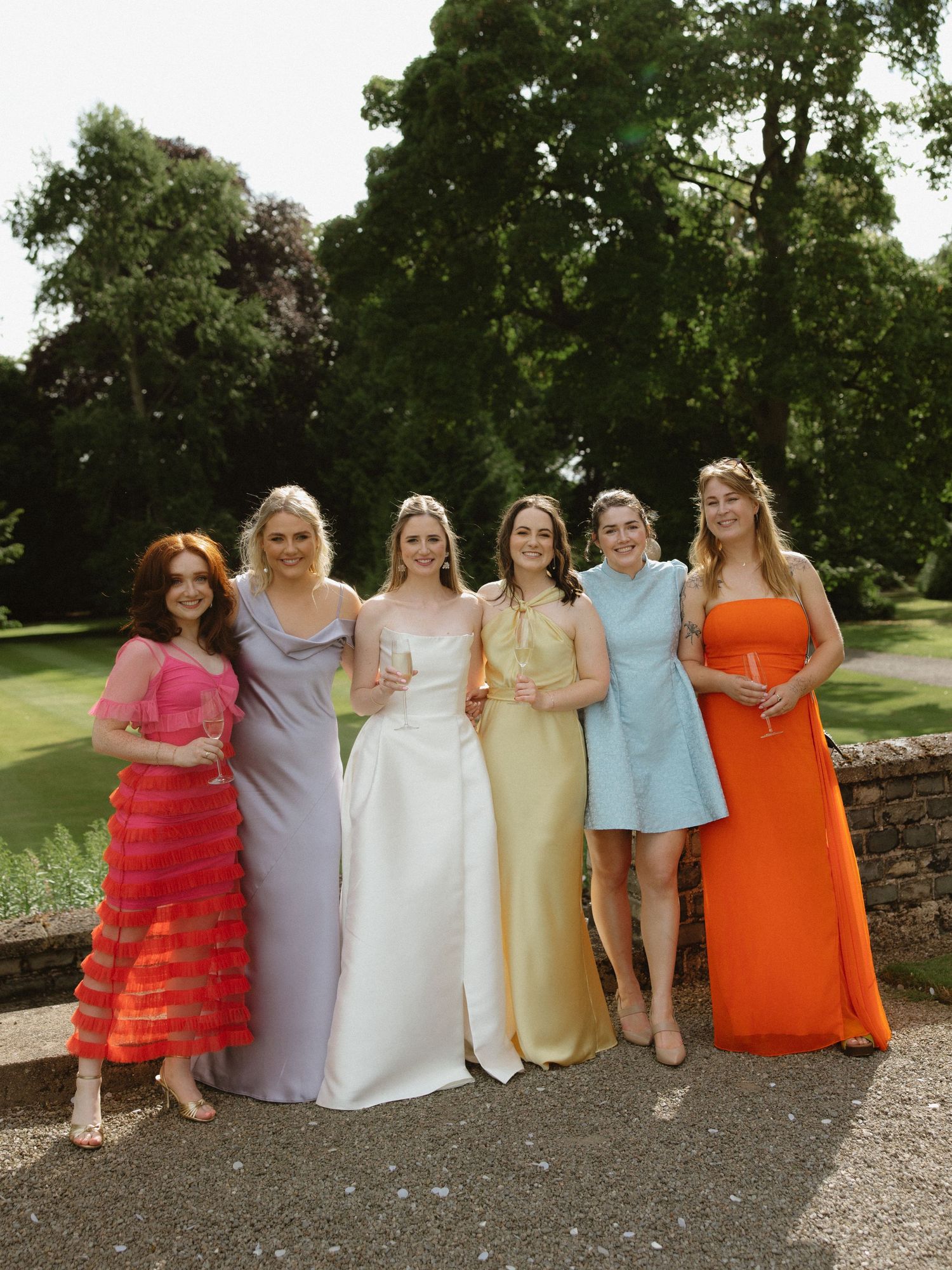 Bridal party in colorful dresses ranging from coral to orange pose together outdoors.