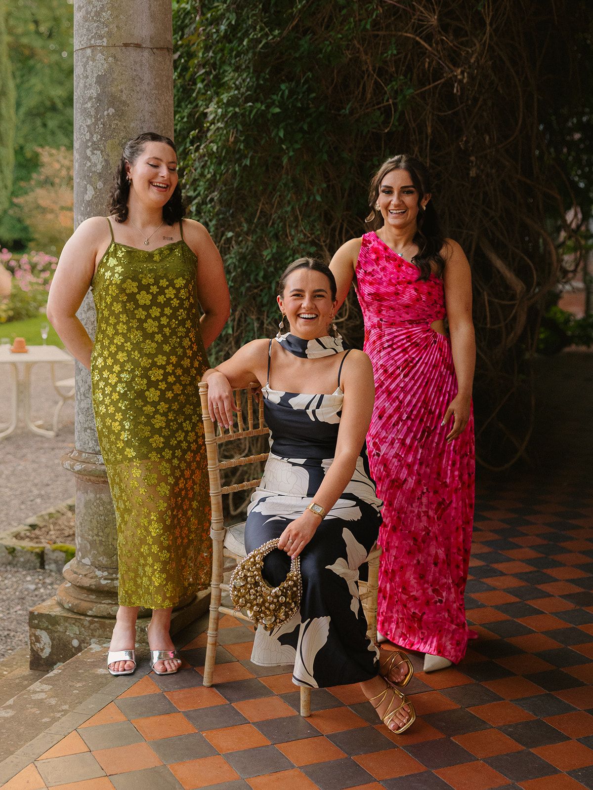 Three friends in colorful summer dresses pose playfully on a tiled patio near garden foliage.