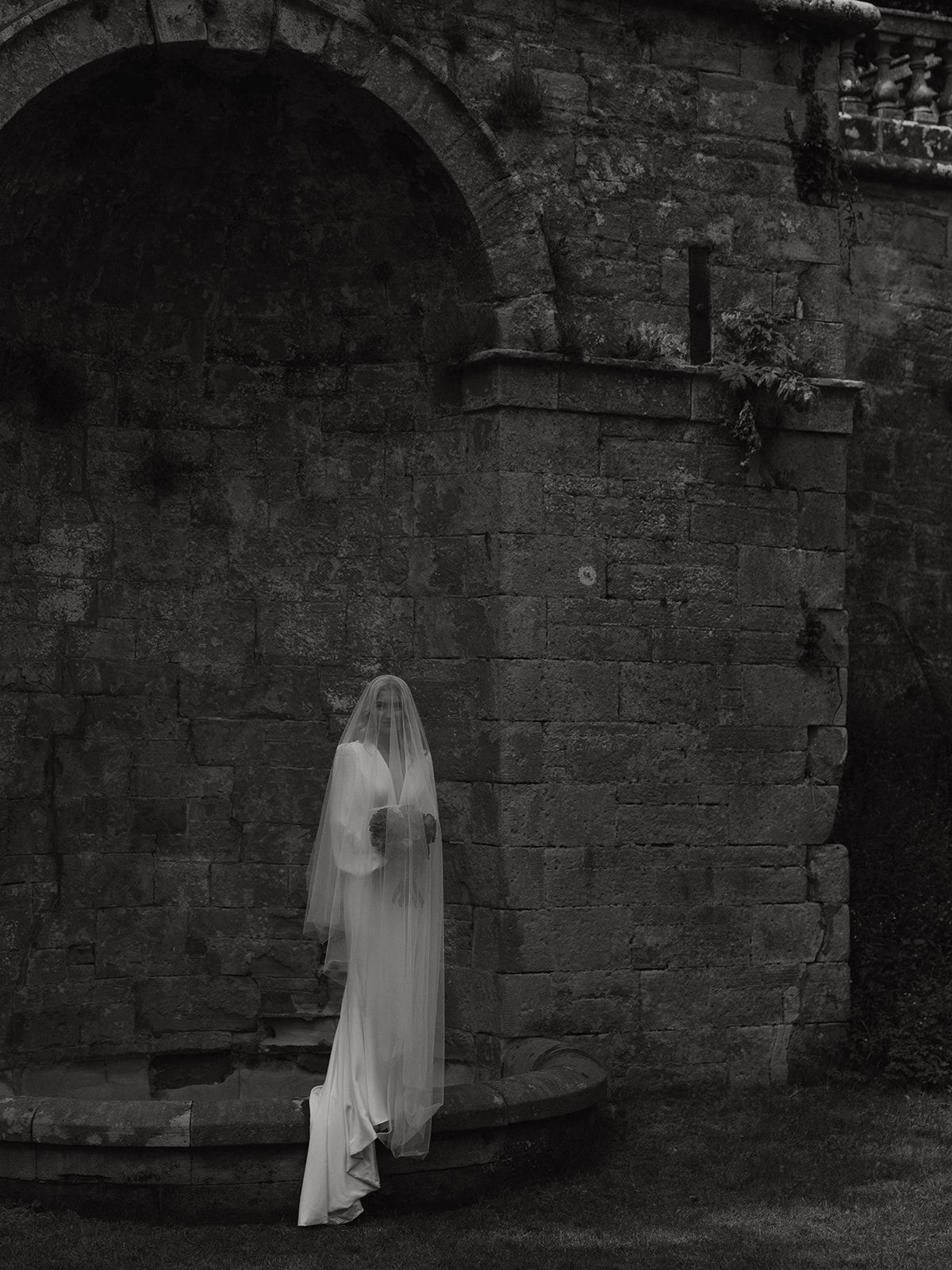 A ghostly figure in a white dress and veil stands beneath a stone archway in a moody black and white scene.