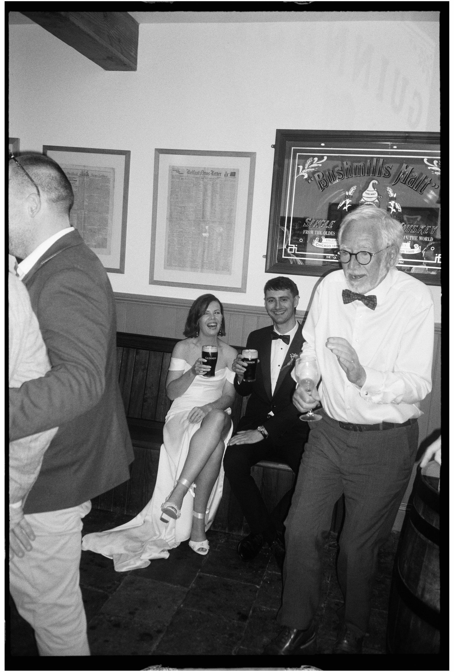 Black and white photo of bride and groom relaxing with guests dancing at a cozy wedding reception.