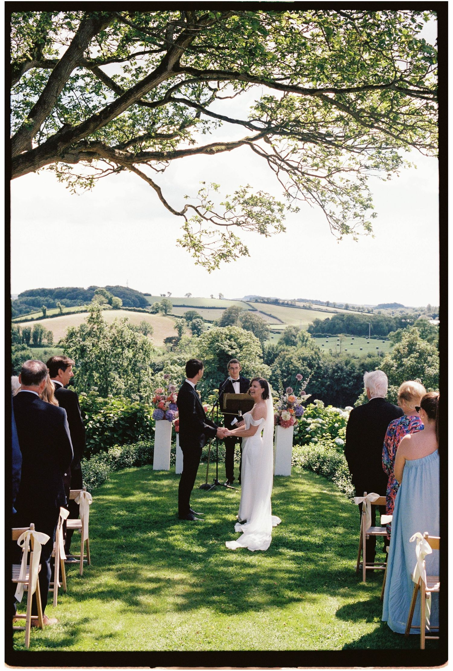 Outdoor wedding ceremony on lush green lawn with rolling hills backdrop, couple exchanging vows under a large tree.