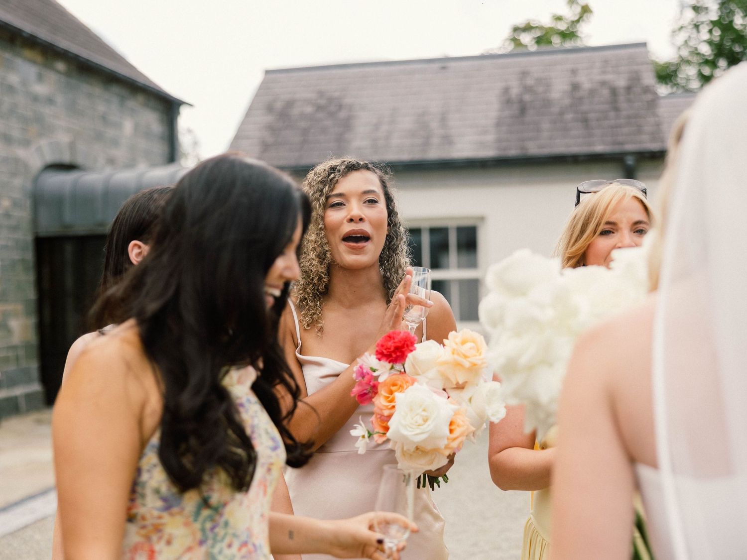 Bridesmaids in pink dresses share joyful moments while holding bouquets outside a stone building.