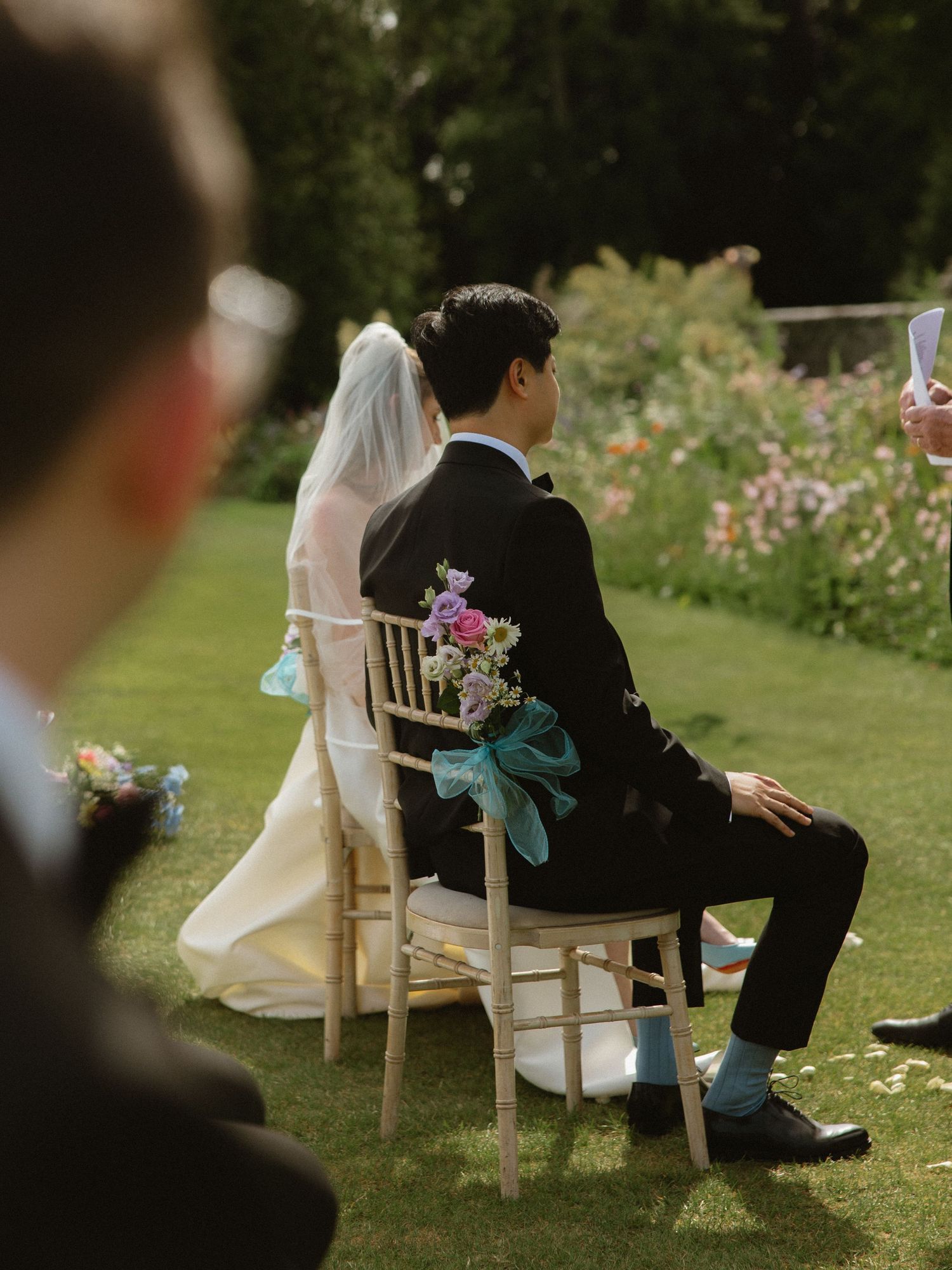 Wedding couple seated on decorated chairs during an outdoor garden ceremony.