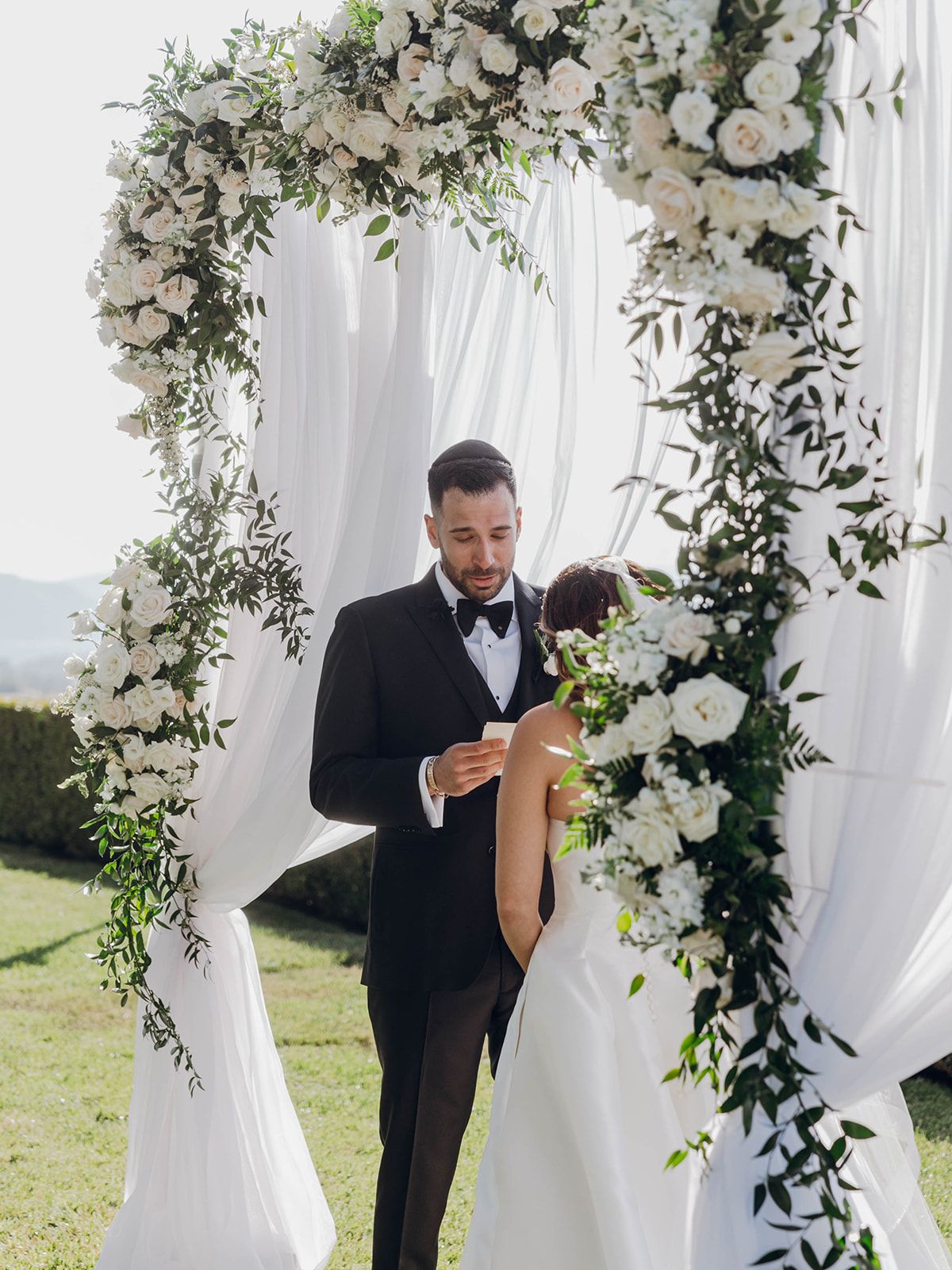 A wedding ceremony setup with an elegant floral arch made of white roses and greenery against flowing white drapery outdoors.