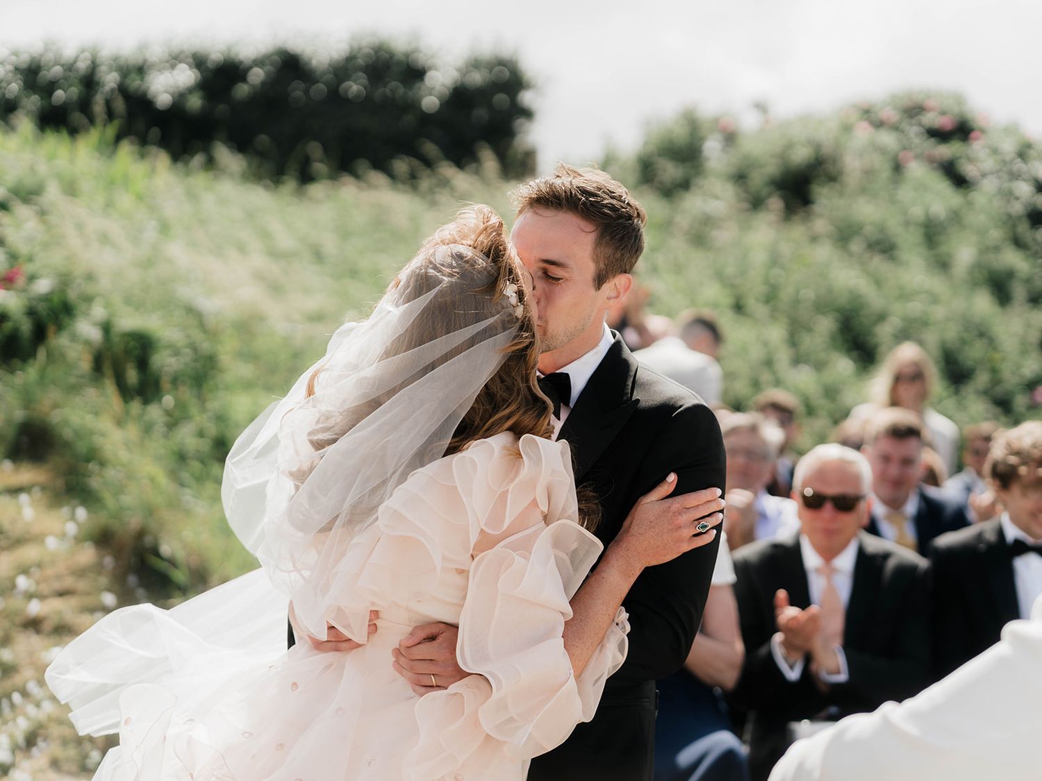 A romantic embrace during an outdoor wedding with guests looking on.