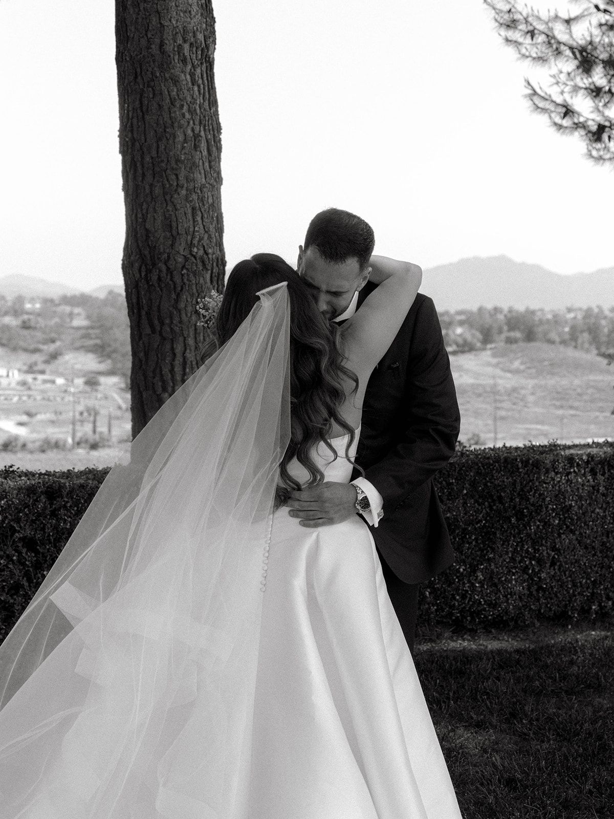 A romantic black and white wedding photo shows a couple embracing beneath a pine tree with a scenic mountain backdrop.