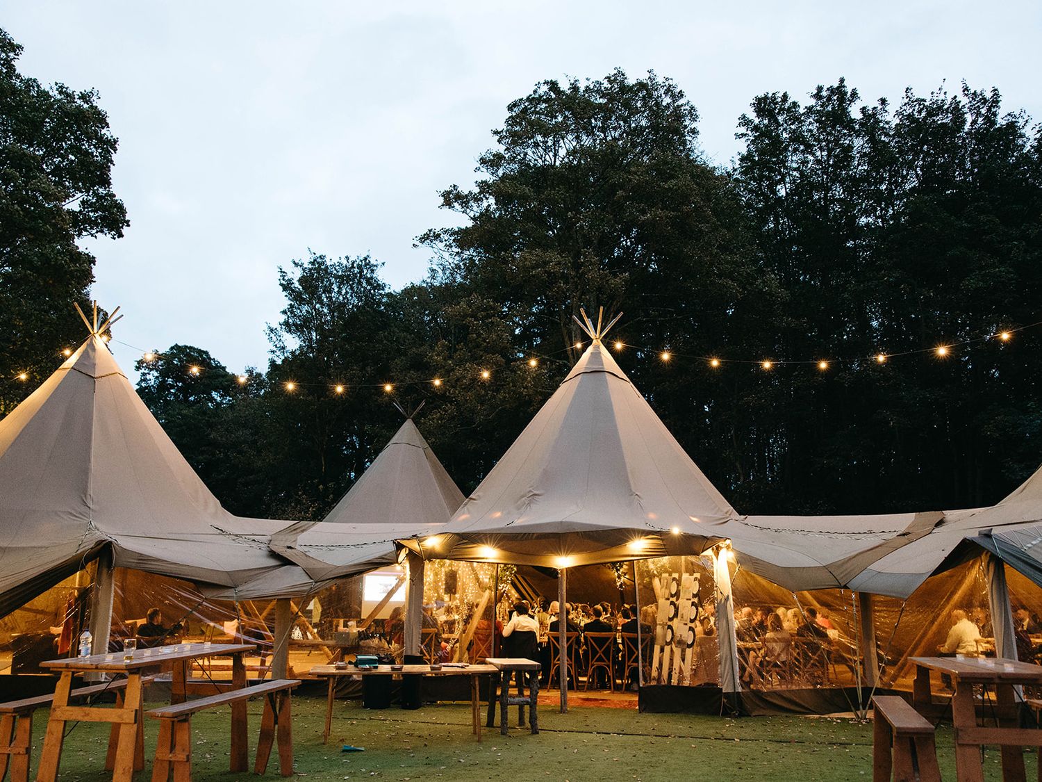 String lights illuminate a cluster of elegant white teepee-style tents at an outdoor event setup at dusk.