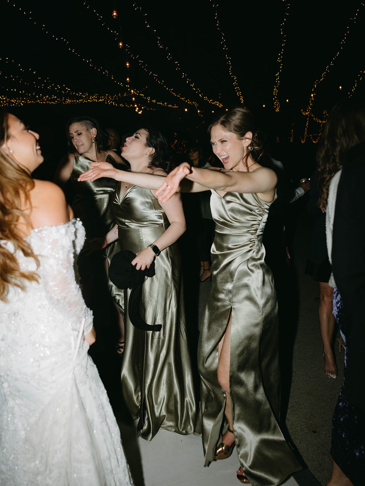 Wedding guests dancing in elegant satin gowns and formal attire under string lights at an evening reception.