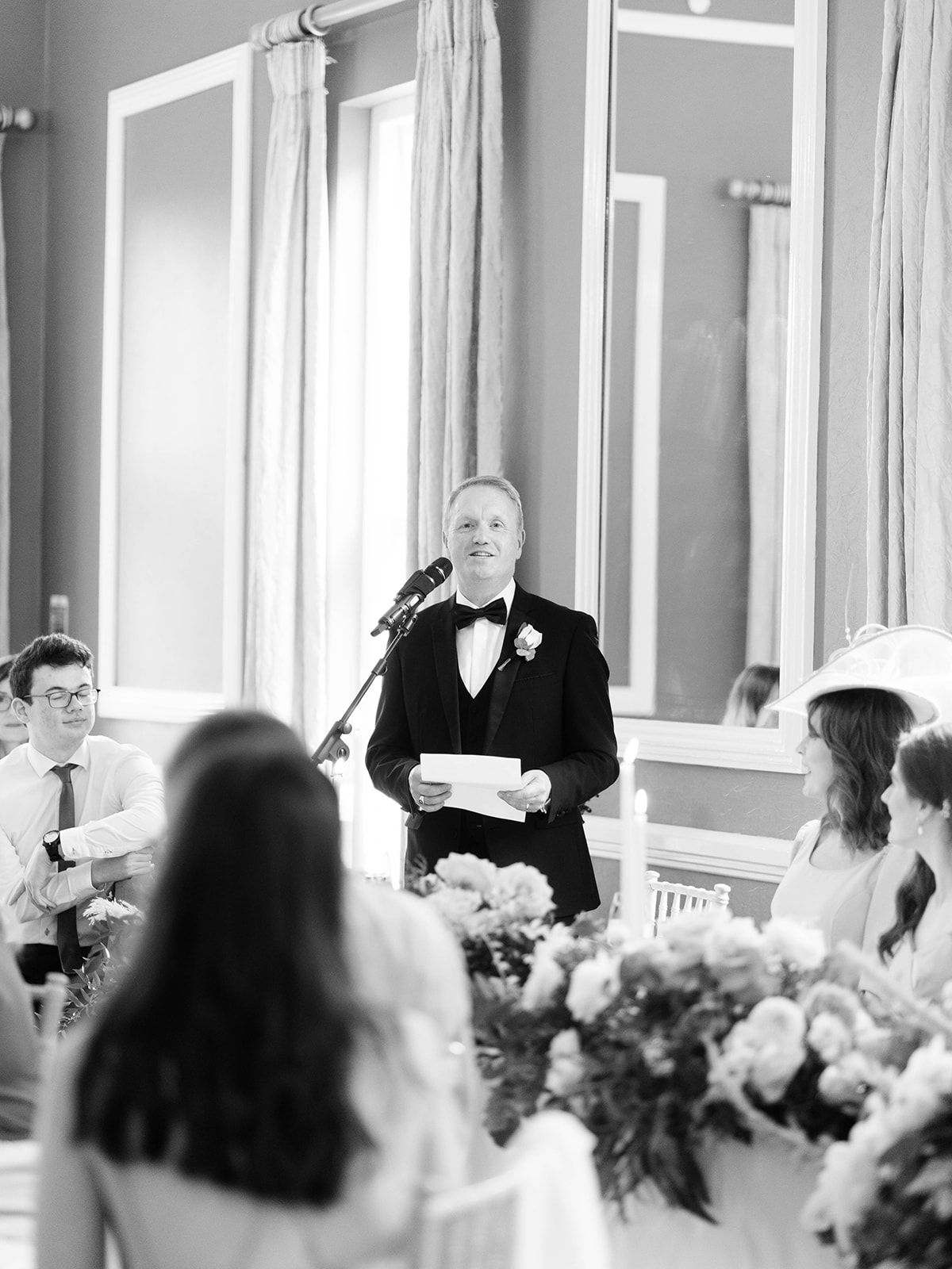 A person in a black tuxedo gives a wedding reception speech while standing at a table decorated with flower arrangements.