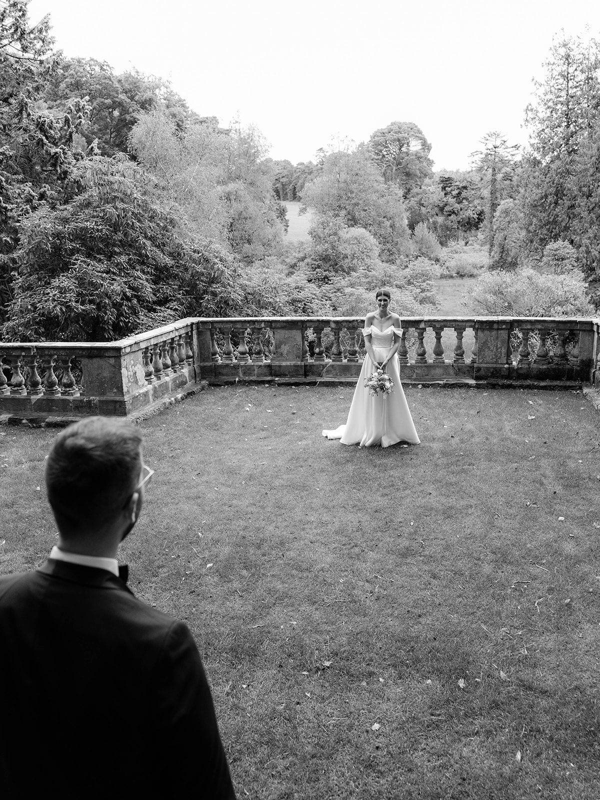 A black and white photograph of two people in formal attire on a garden terrace with ornate stone balustrades and trees.