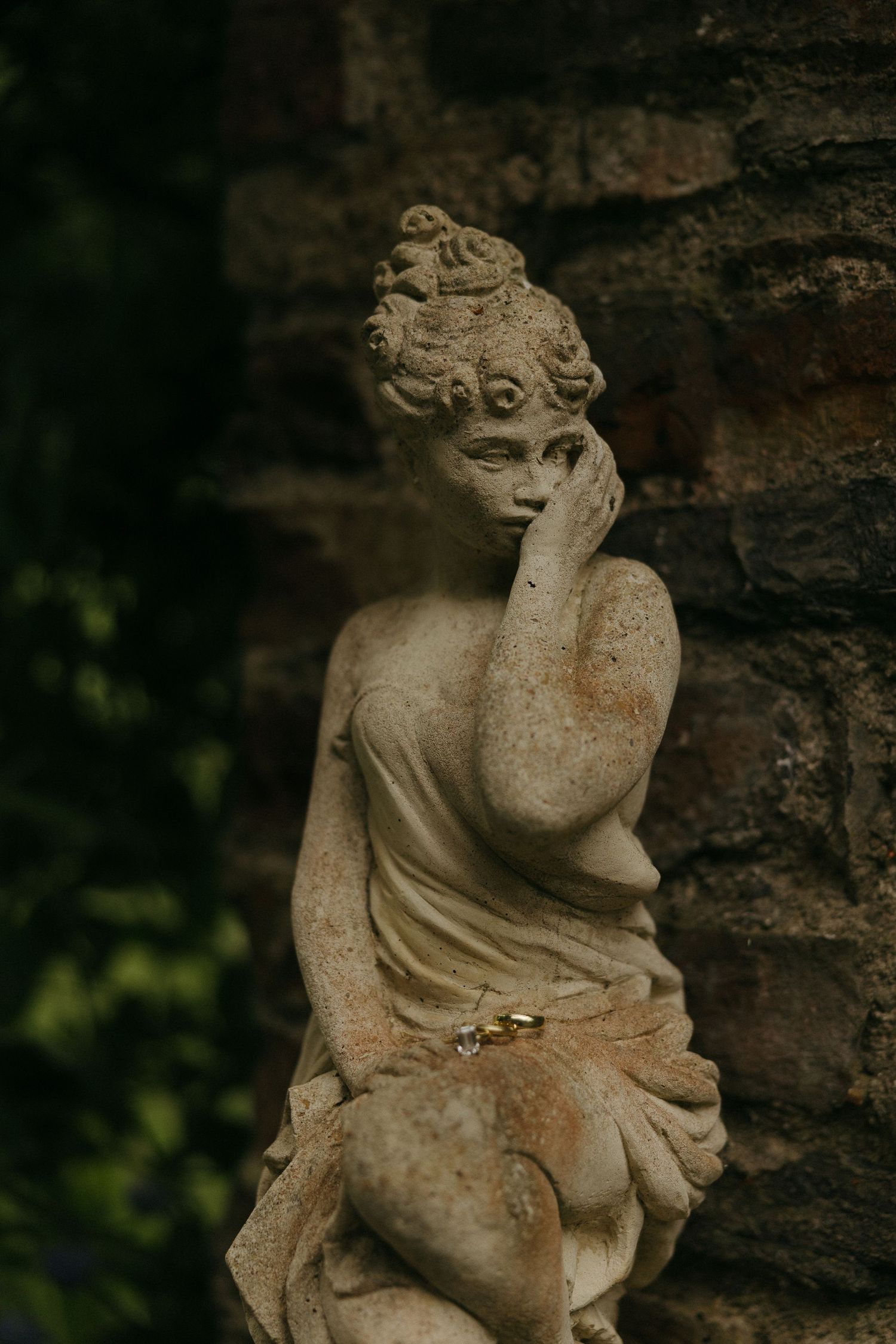 Stone garden statue of a contemplative woman in classical dress, resting her chin on her hand against a rustic brick wall.
