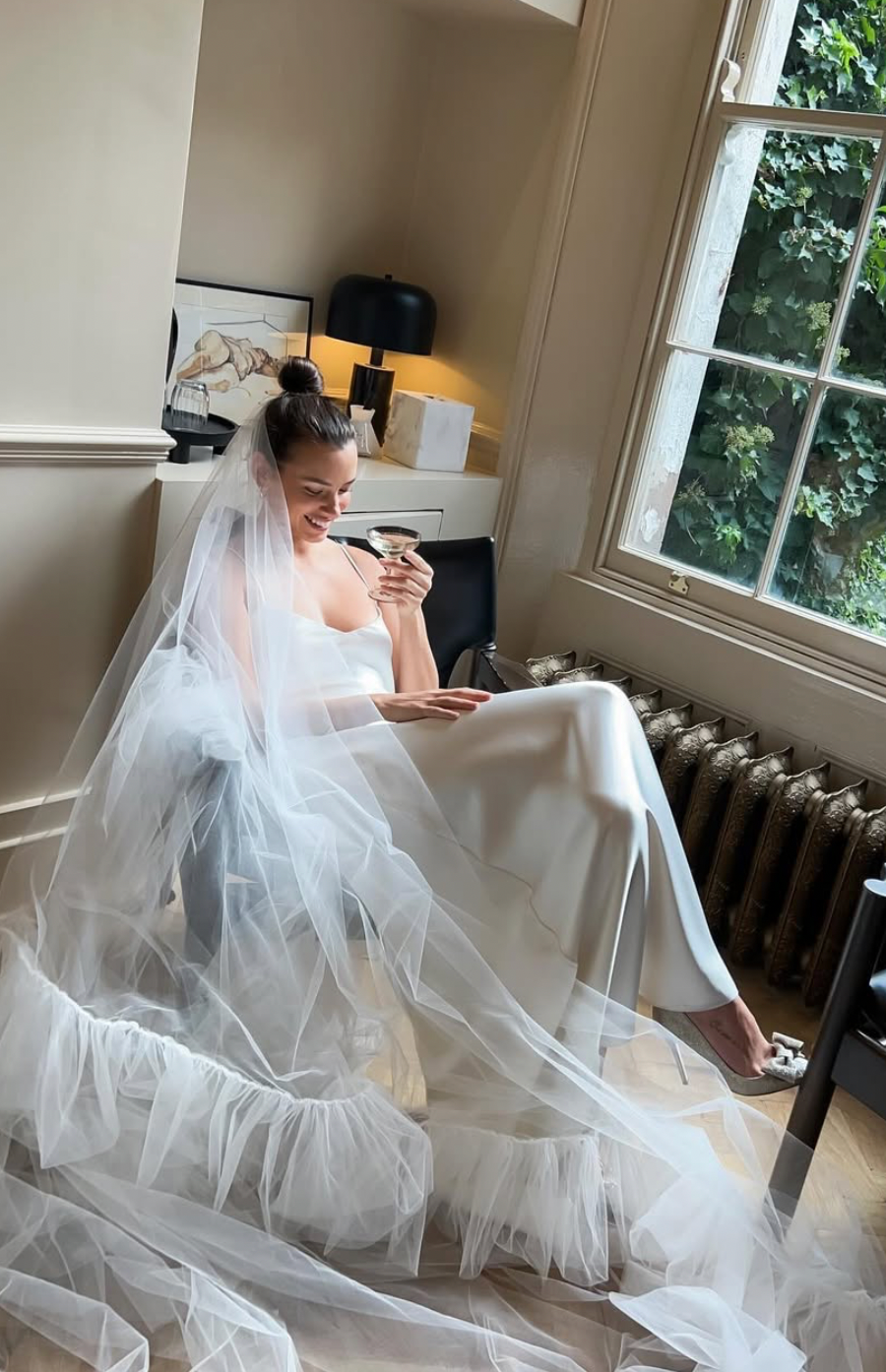 A bride in a flowing white wedding dress and veil sits in a sunlit room reading a book near a window.