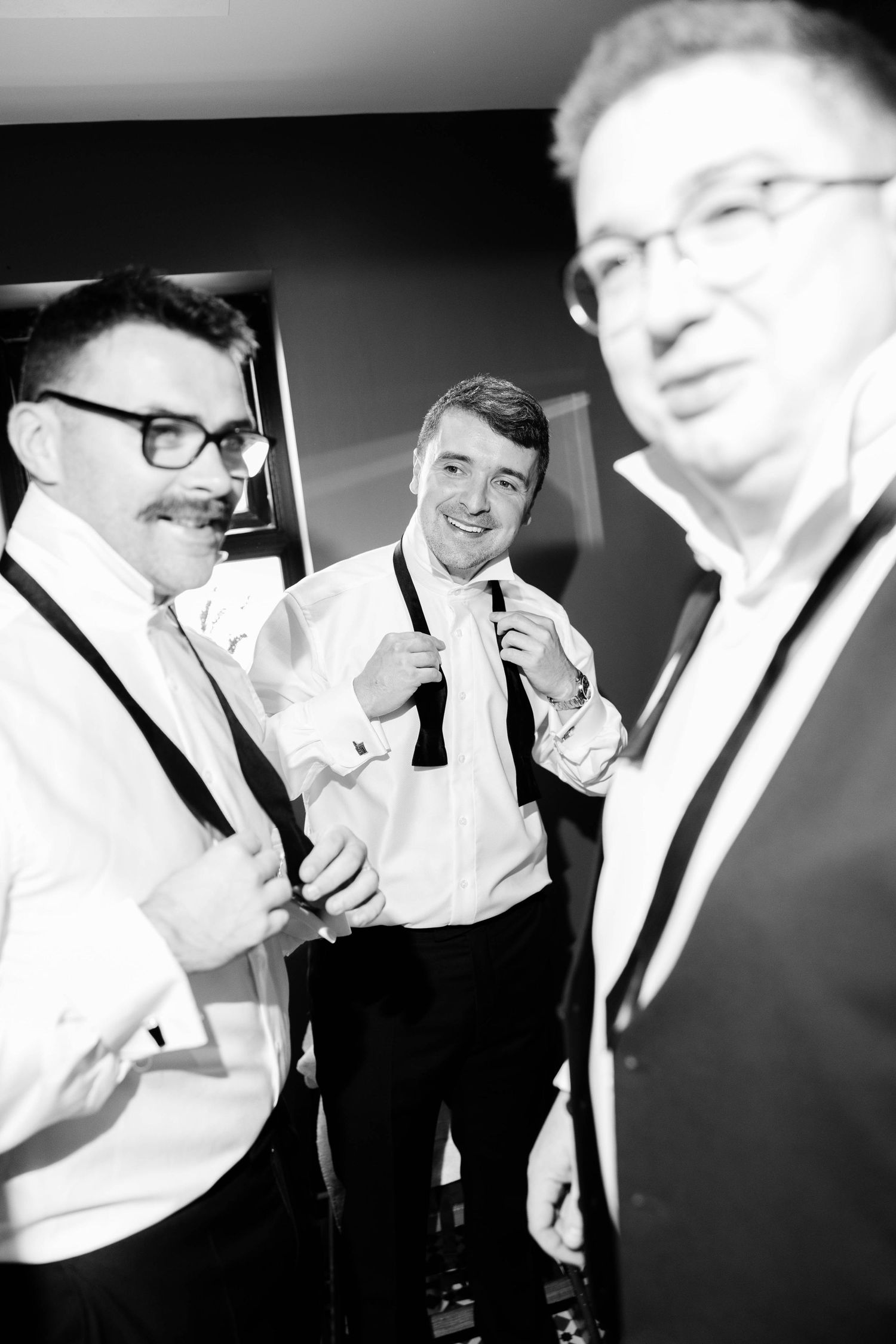 Three men in formal attire share a candid black and white moment at what appears to be a wedding or formal event.