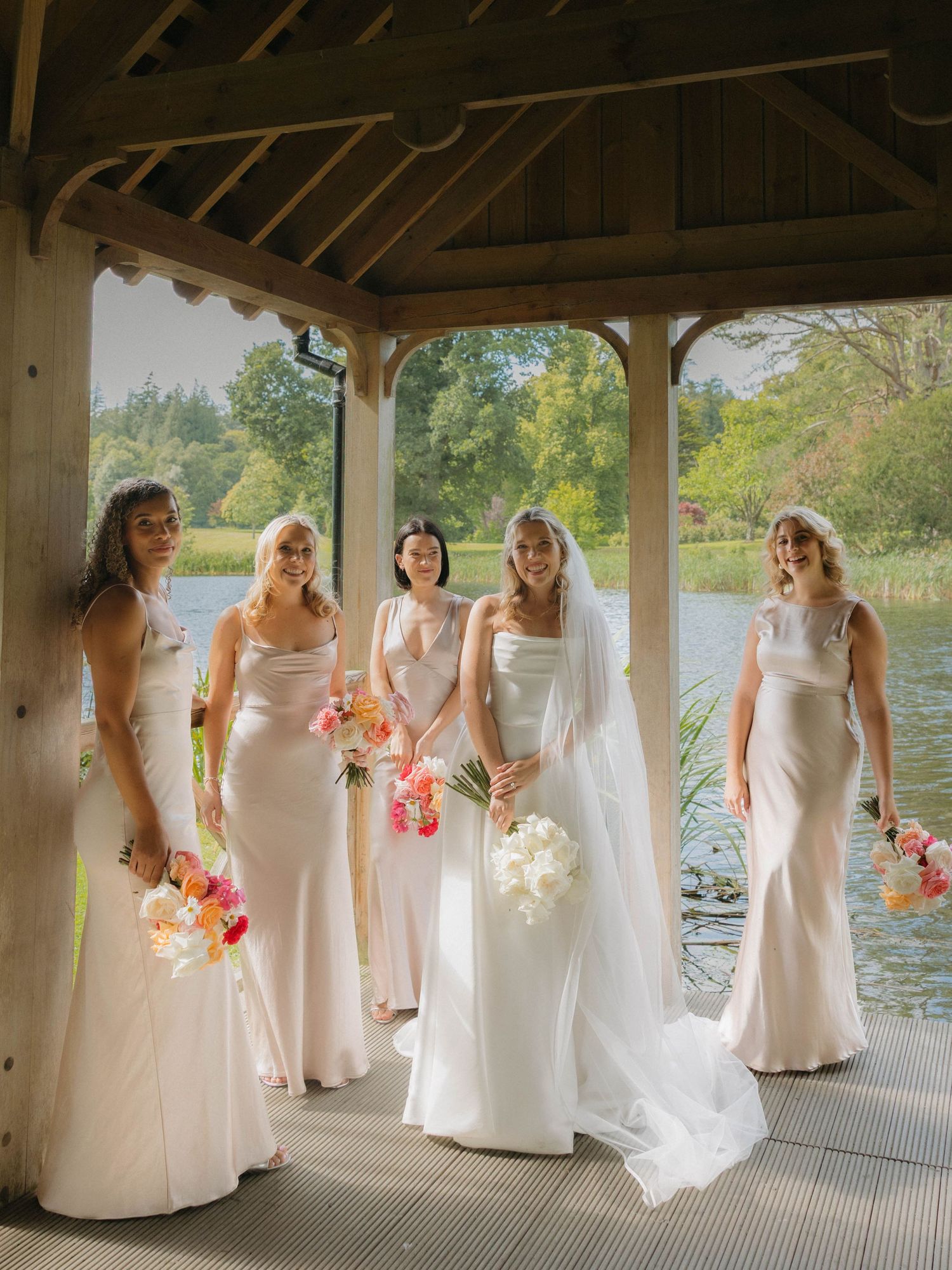 Bridal party in elegant white dresses poses with bouquets in a covered dock by the water.