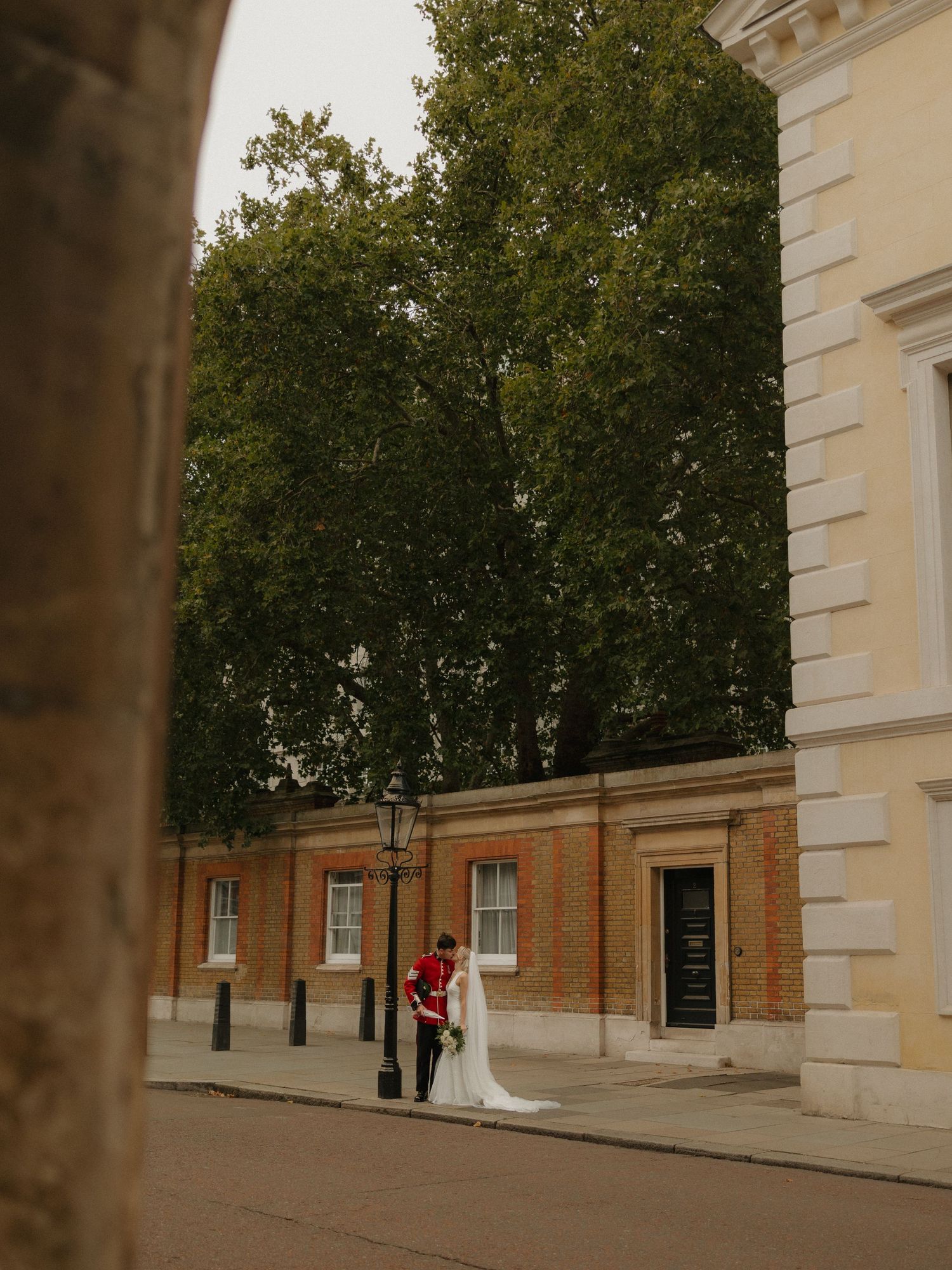 Couple embraces in front of historic brick building with tall trees and white architectural details in Greenwich, London.