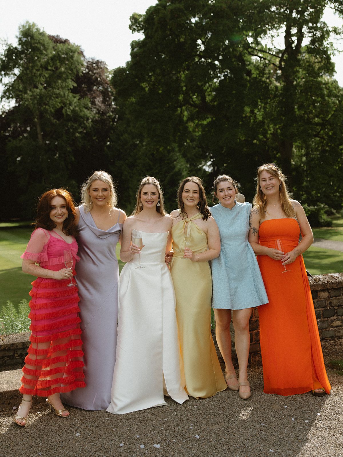 A group poses outdoors wearing colorful formal dresses in pink, lavender, white, yellow, blue and orange against a green garden.