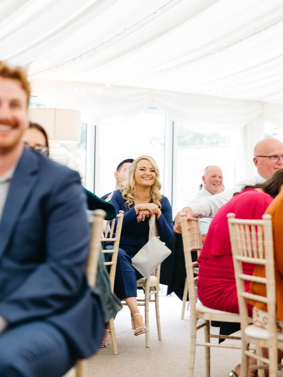Guests seated in white Chiavari chairs laugh during a wedding reception in a bright tent-covered venue.