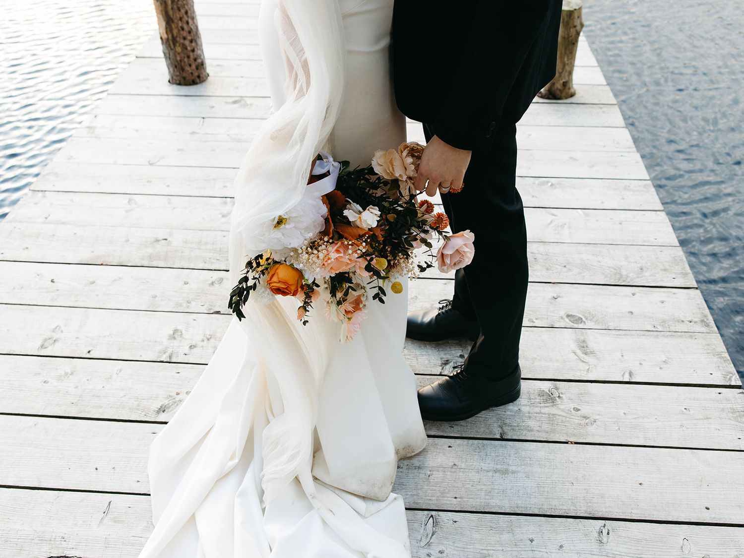 A couple in wedding attire stands on a white dock holding a vibrant fall-colored bouquet of flowers.