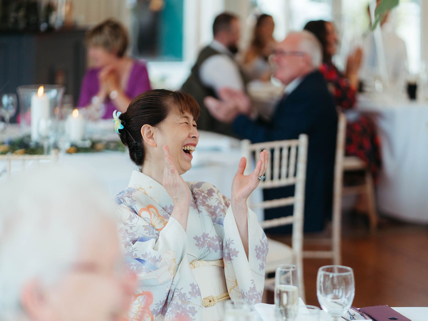 A dinner guest wearing a floral kimono laughs joyfully while seated at a reception table in a bright dining room.