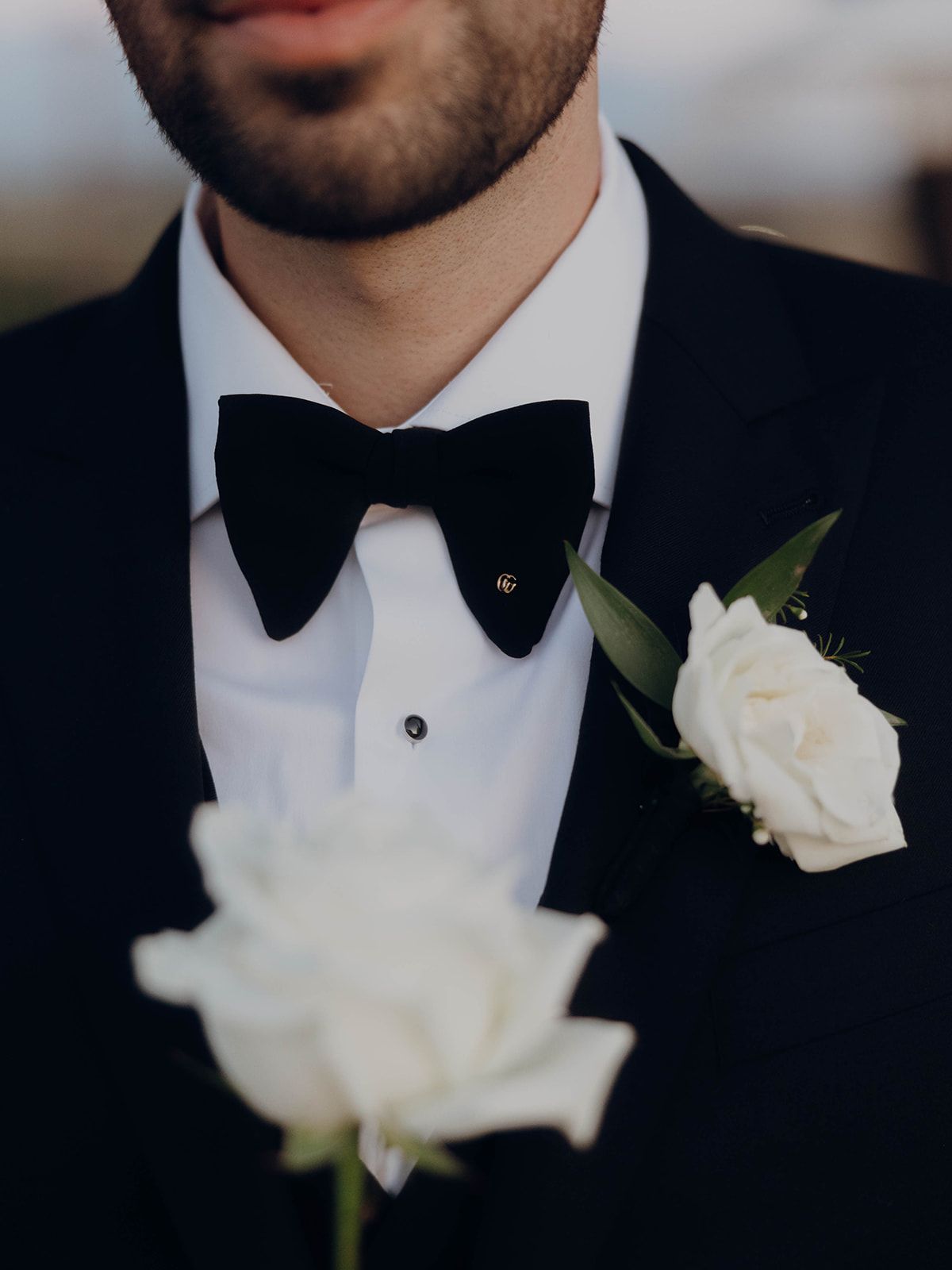 Elegant groom's formal attire with black bow tie, white boutonniere flower, and black tuxedo jacket in classic style.