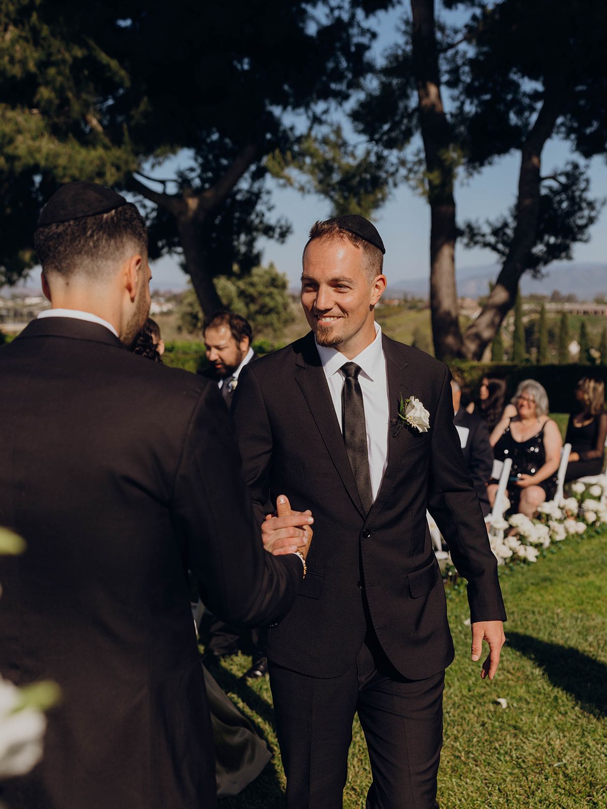 Two people in dark suits exchanging greetings at an outdoor wedding ceremony with guests seated in background.