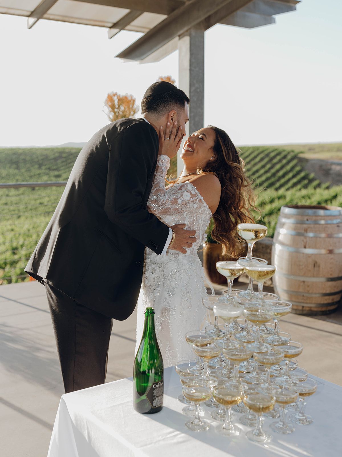A romantic moment between newlyweds at a vineyard with a champagne tower and rustic wine barrels in the setting sun.