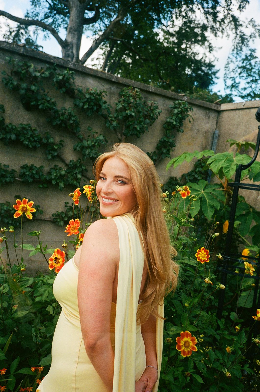 Person in yellow dress poses in garden with blooming orange lantana flowers and stone wall backdrop.