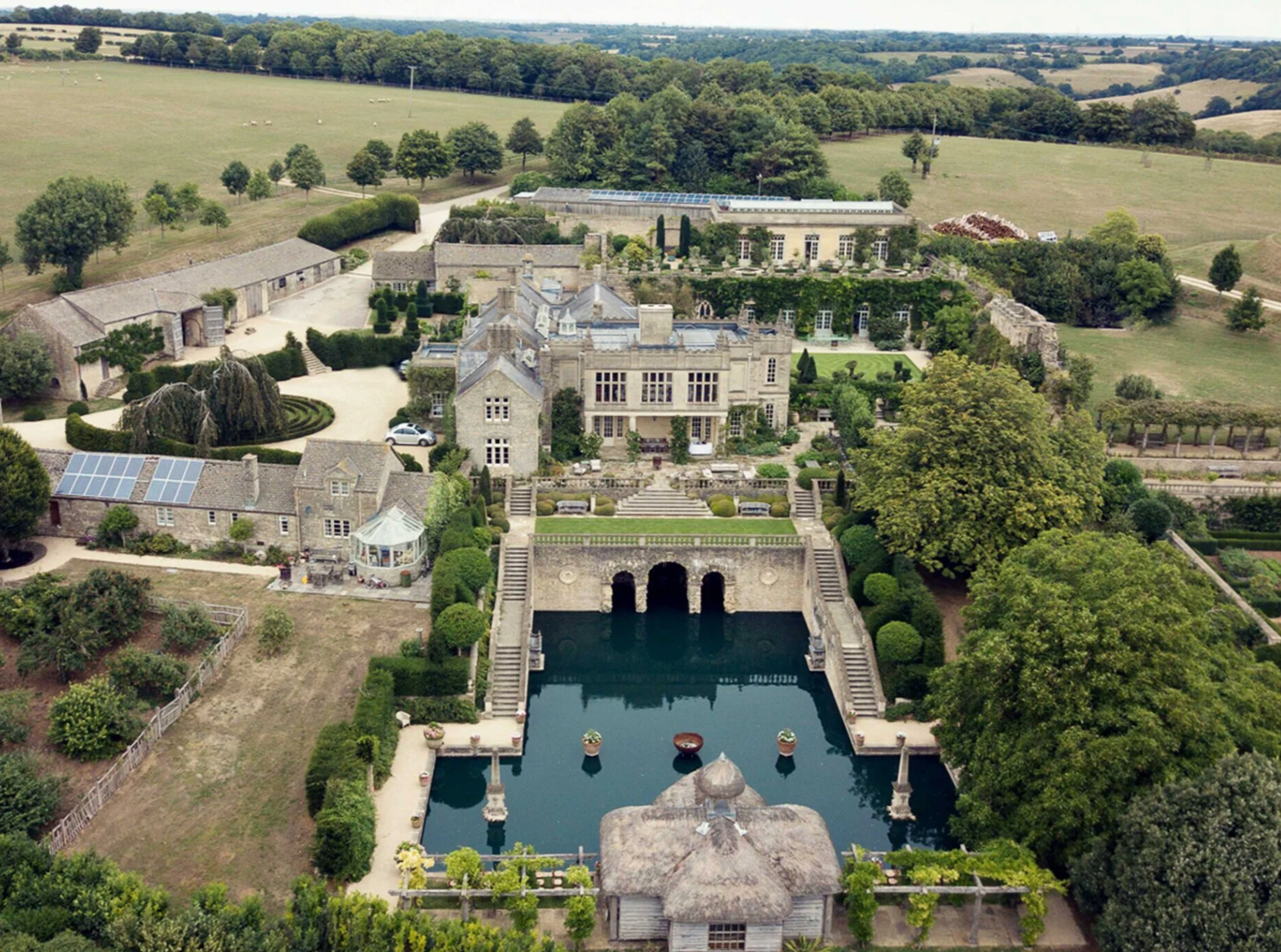 Aerial view of a grand English manor house with formal gardens and a large reflecting pool surrounded by manicured grounds.