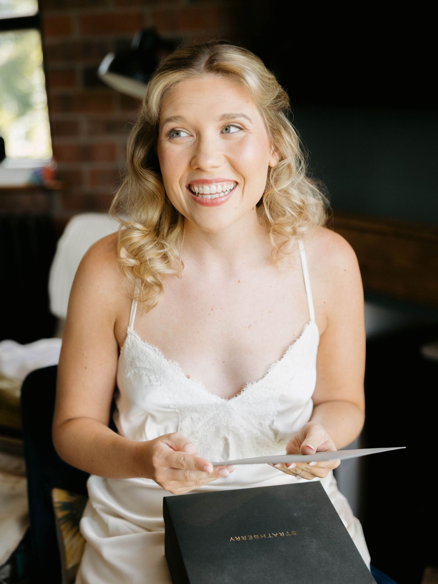 A person in a white dress smiles warmly while holding papers in an indoor setting with natural lighting.