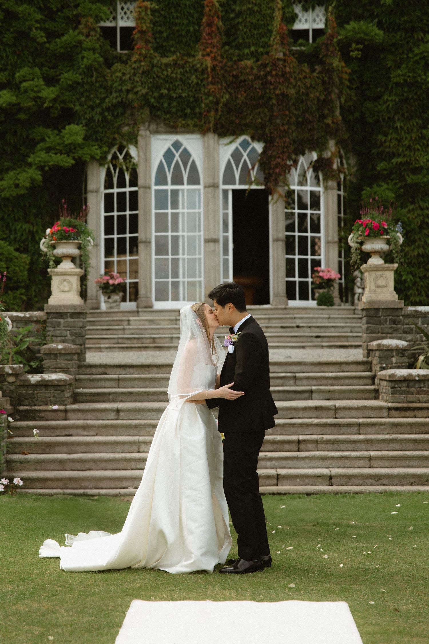 A couple in wedding attire shares a tender moment on the lawn steps of an elegant stone building with Gothic windows.