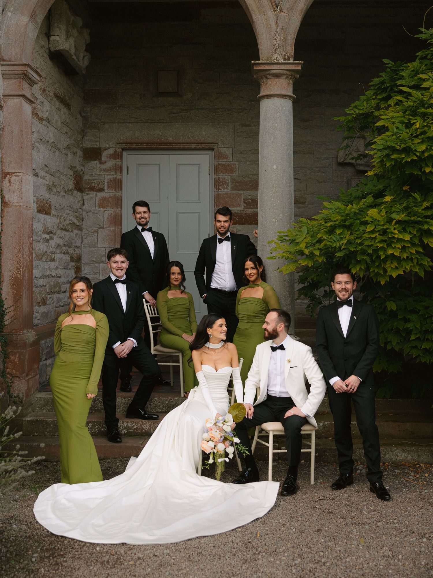 Wedding party in olive green and formal attire poses in rustic stone courtyard setting.