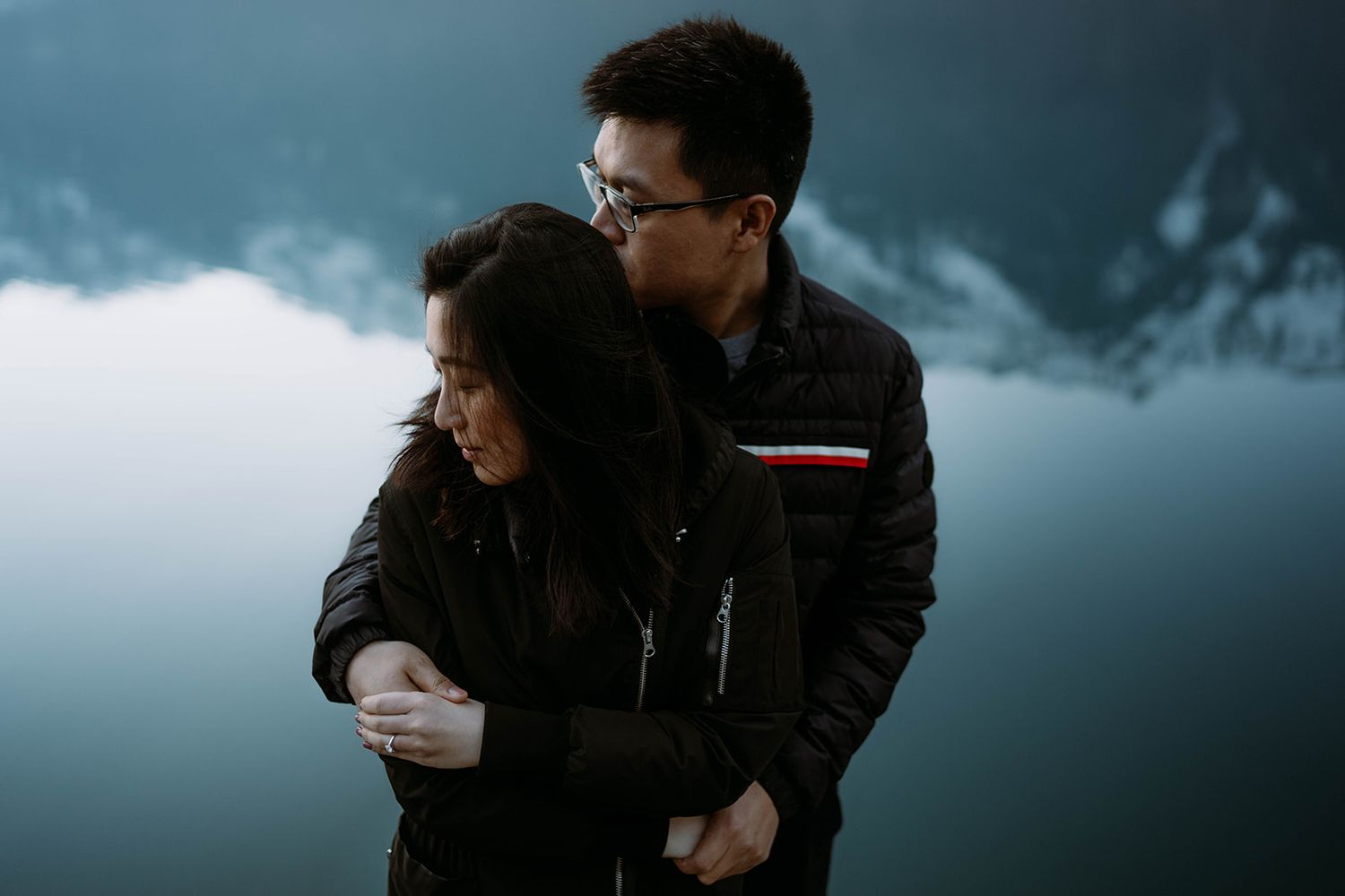 Couple embracing tenderly by a misty mountain lake, wearing dark jackets in a moody, romantic outdoor setting.