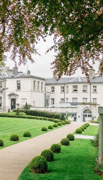 Elegant white Georgian manor house with manicured topiary gardens and gravel pathway surrounded by lush green lawns.