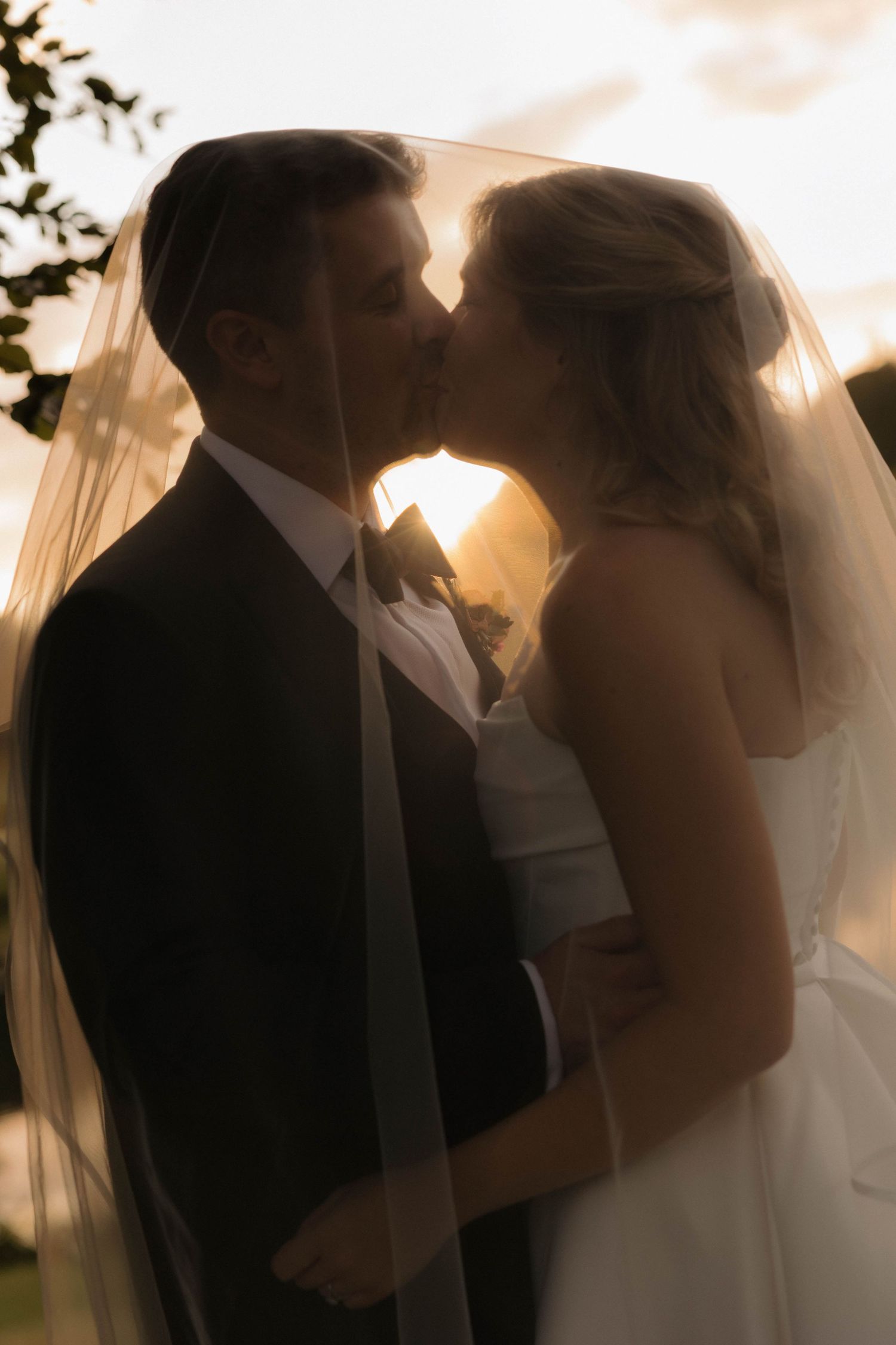 Silhouetted wedding couple embracing during golden hour with a delicate veil floating in the breeze.