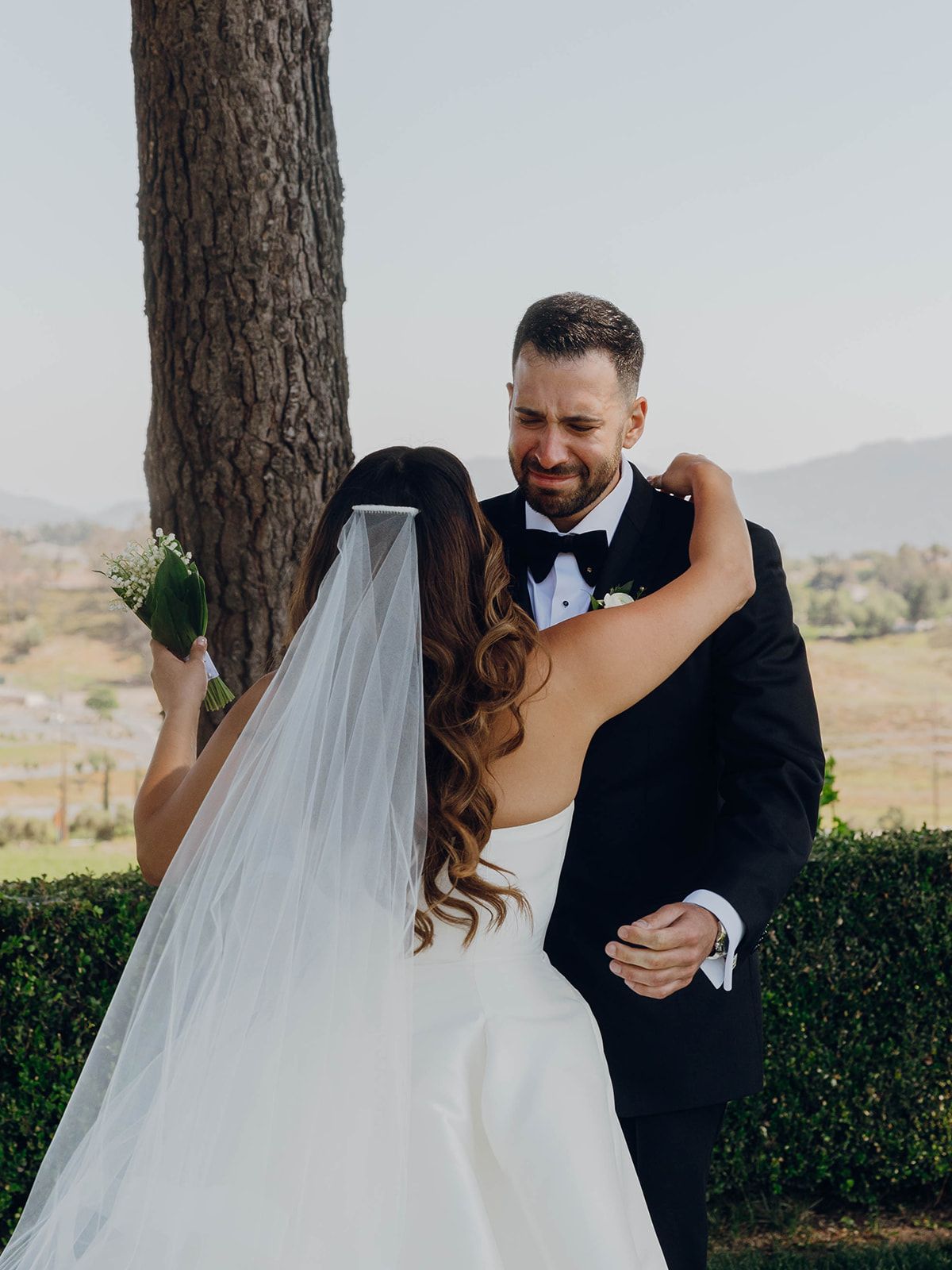 A couple shares a sweet embrace at their outdoor wedding with mountains in the backdrop and a large tree in the foreground.