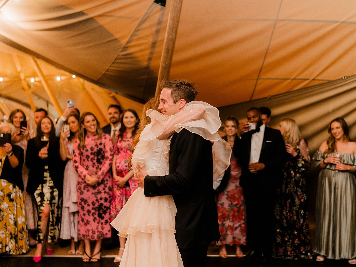 Couple shares their first dance surrounded by guests in floral dresses at tent reception.