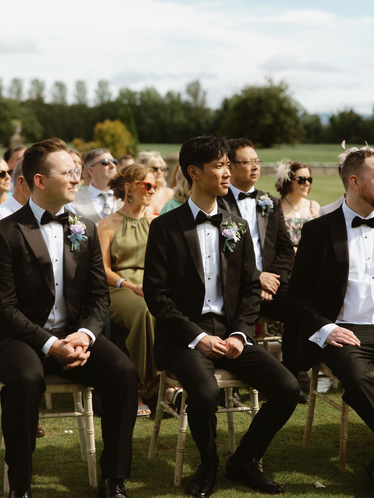 A row of groomsmen in black tuxedos seated during an outdoor wedding ceremony.