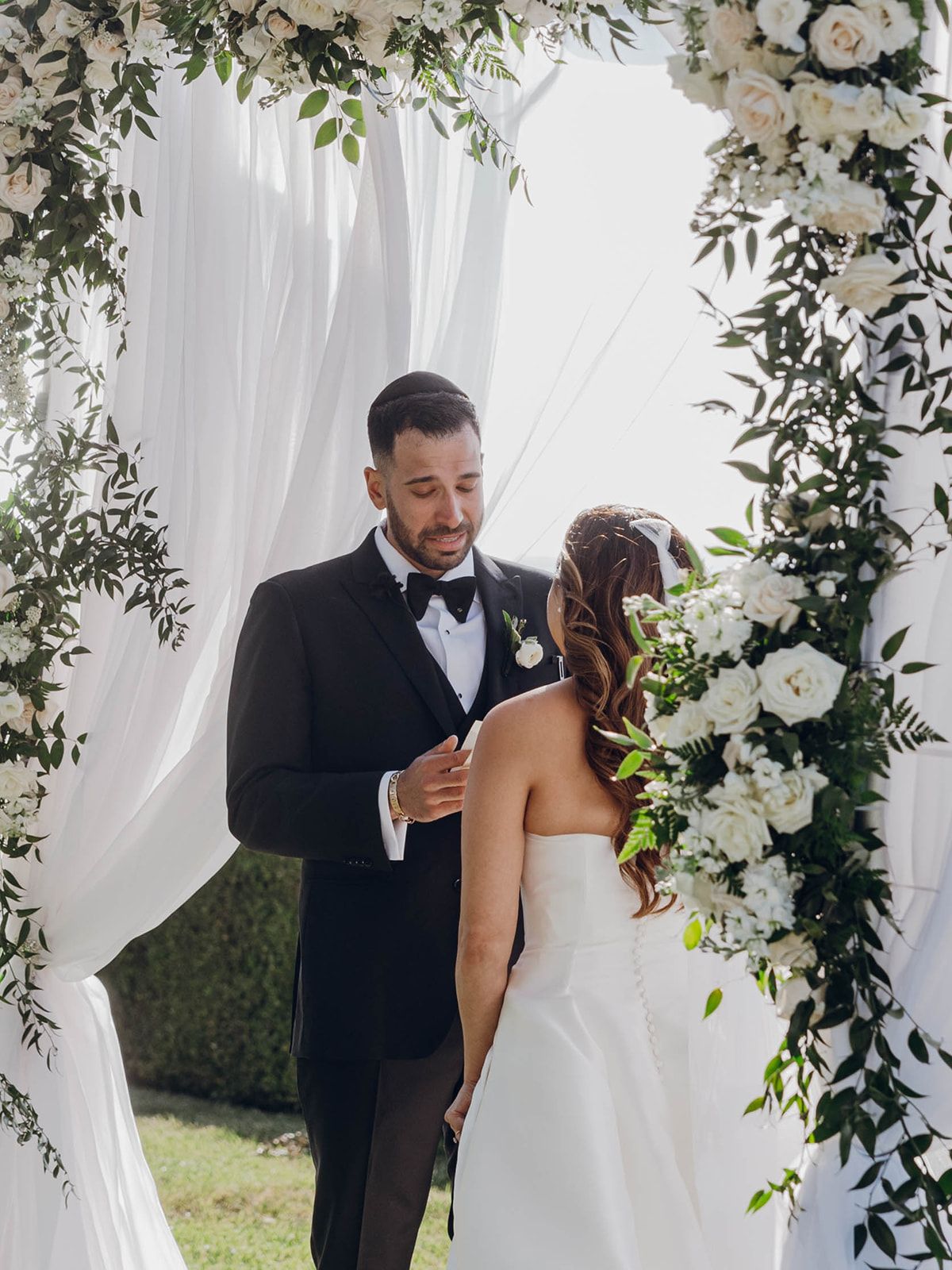A romantic wedding ceremony under a floral arch with white drapery and greenery decorations during an outdoor celebration.