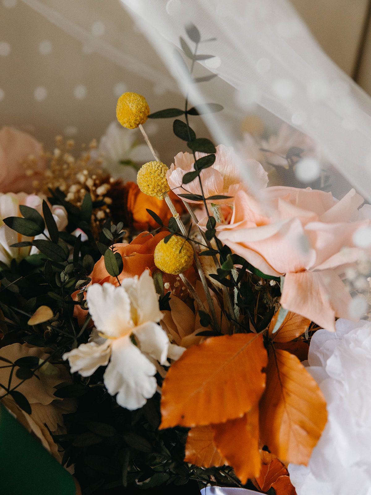 A romantic fall floral arrangement with peach roses, yellow craspedia, and orange autumn leaves against soft beige textiles.