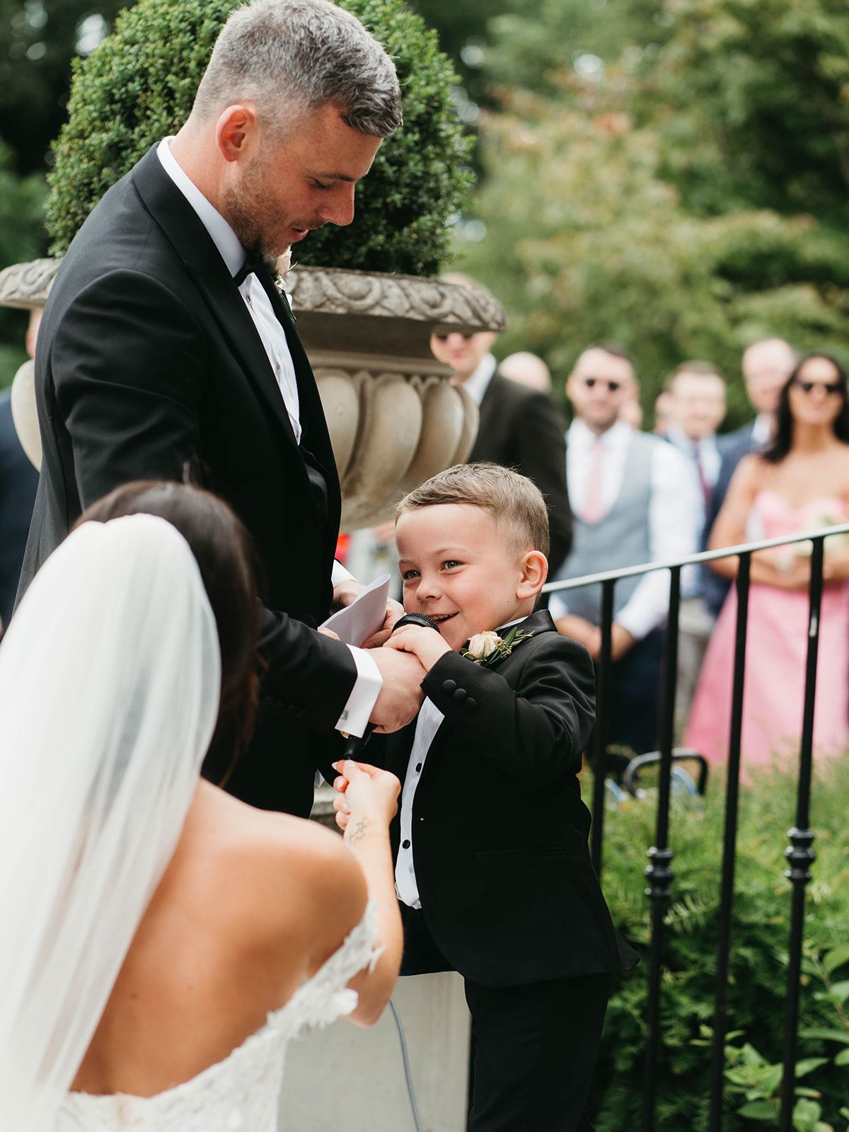 A sweet moment at a wedding ceremony shows a small boy in a black tuxedo sharing a touching interaction with guests.