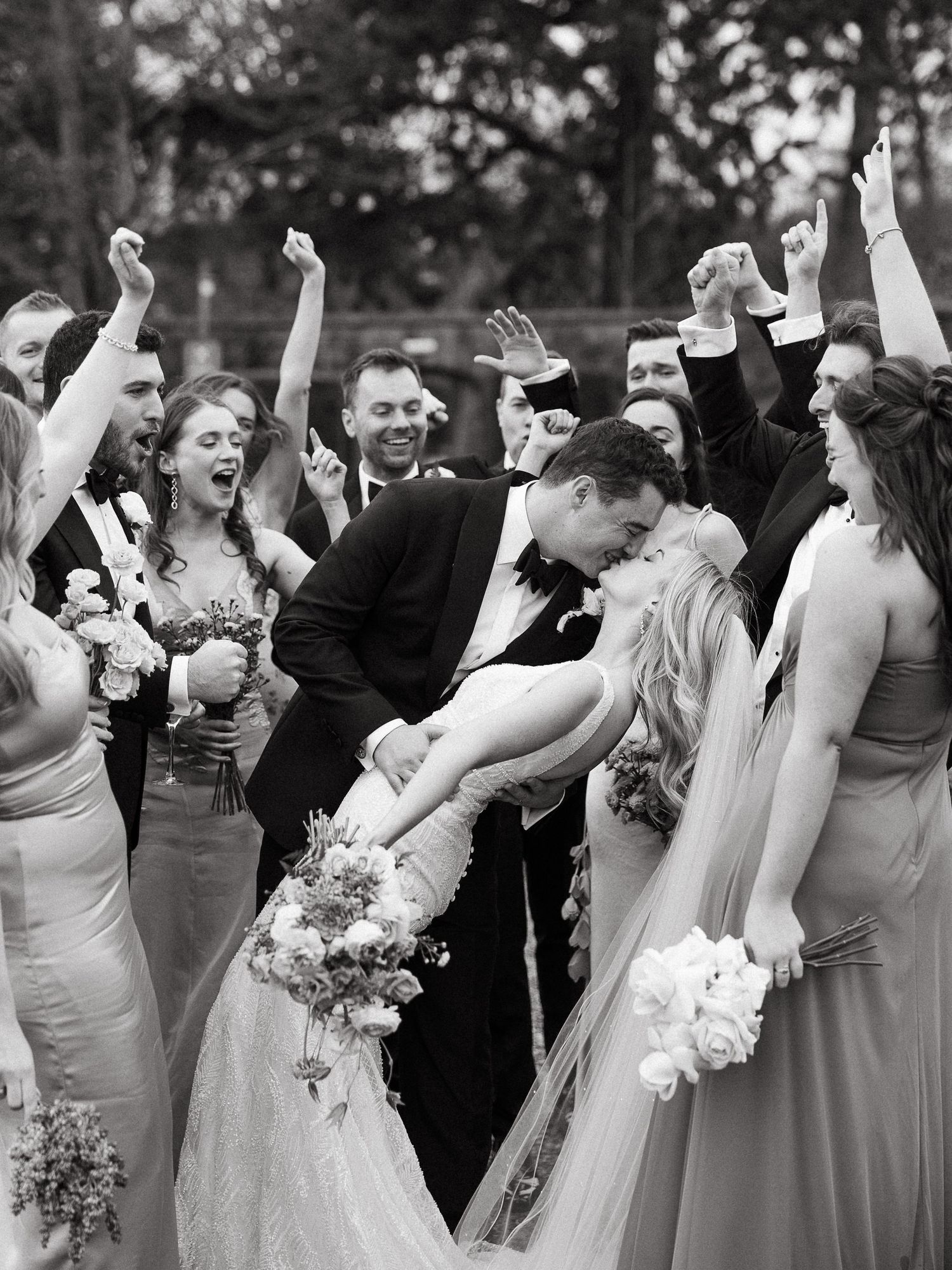 Wedding party celebrates joyfully as couple shares romantic dip kiss during outdoor black and white ceremony.