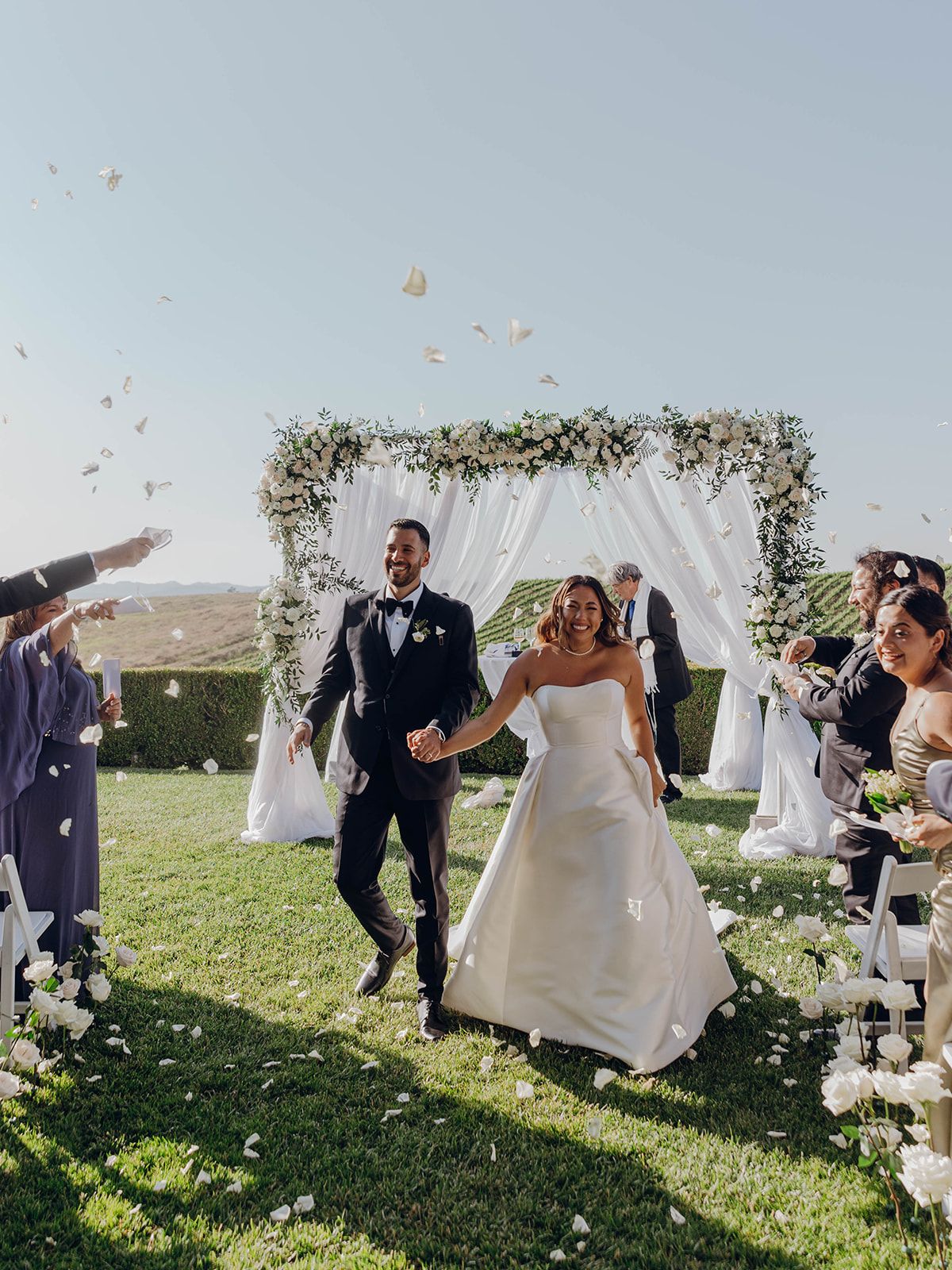 A joyful newlywed couple walks back down the aisle under flower petals at their outdoor garden wedding ceremony.