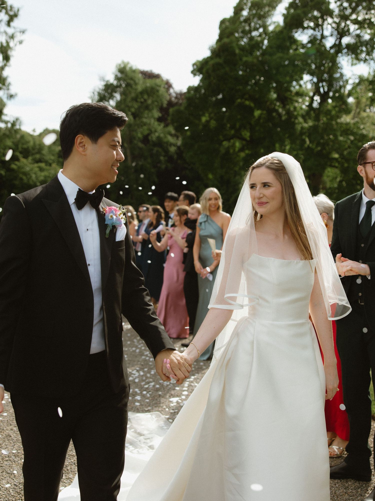 A joyful wedding ceremony with guests tossing confetti at the couple on a tree-lined path.